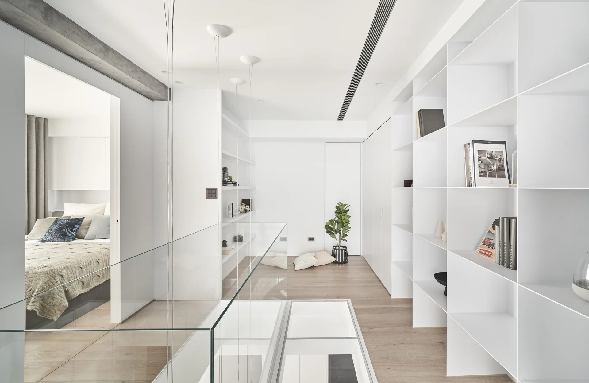 A bright minimalist loft landing features built-in white grid shelving, light wood floors, a glass floor insert, and a transparent safety railing.