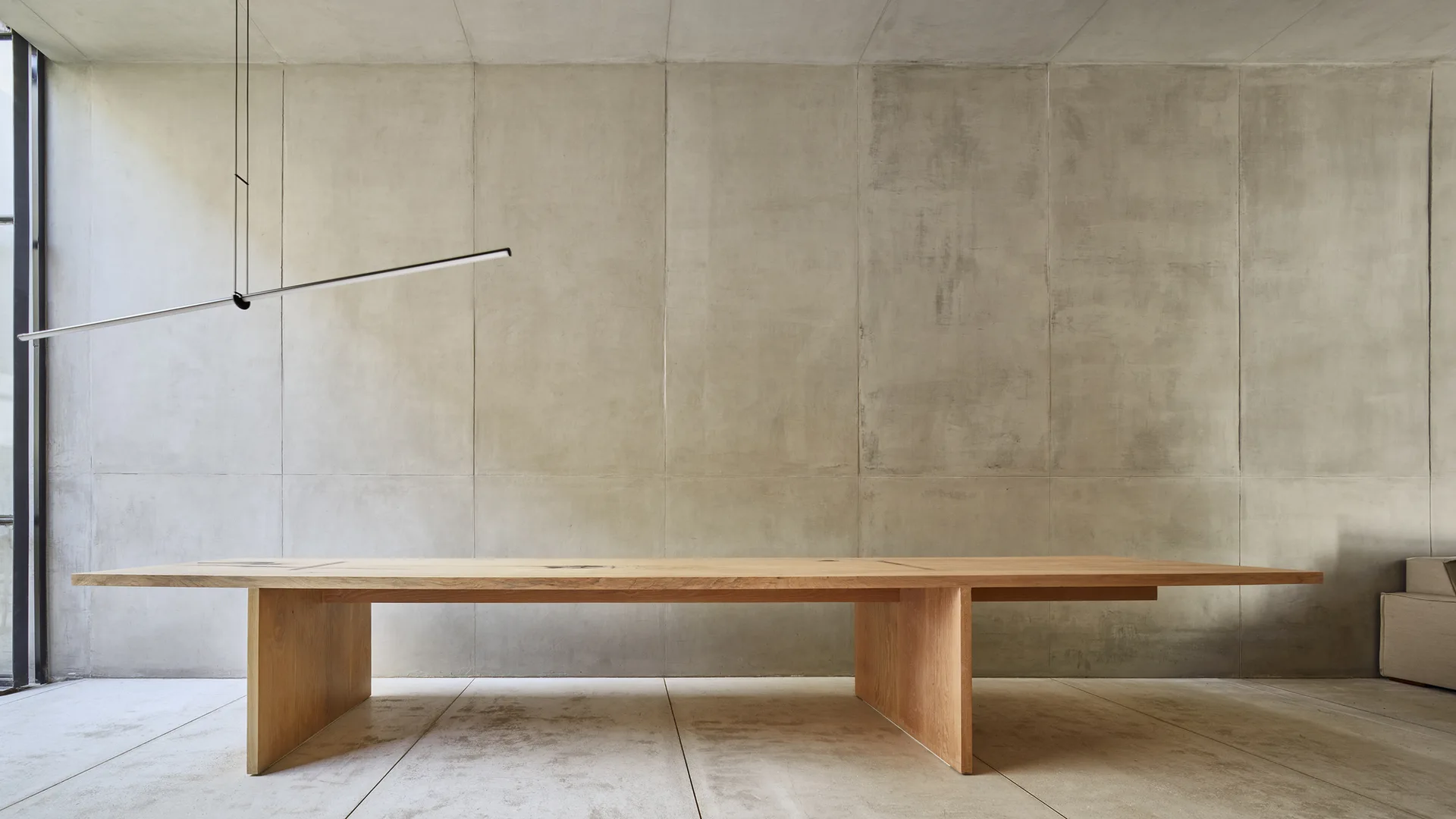 A long, light wood dining table sits in a minimalist room with concrete panel walls, concrete tile flooring, and a thin linear black pendant light.