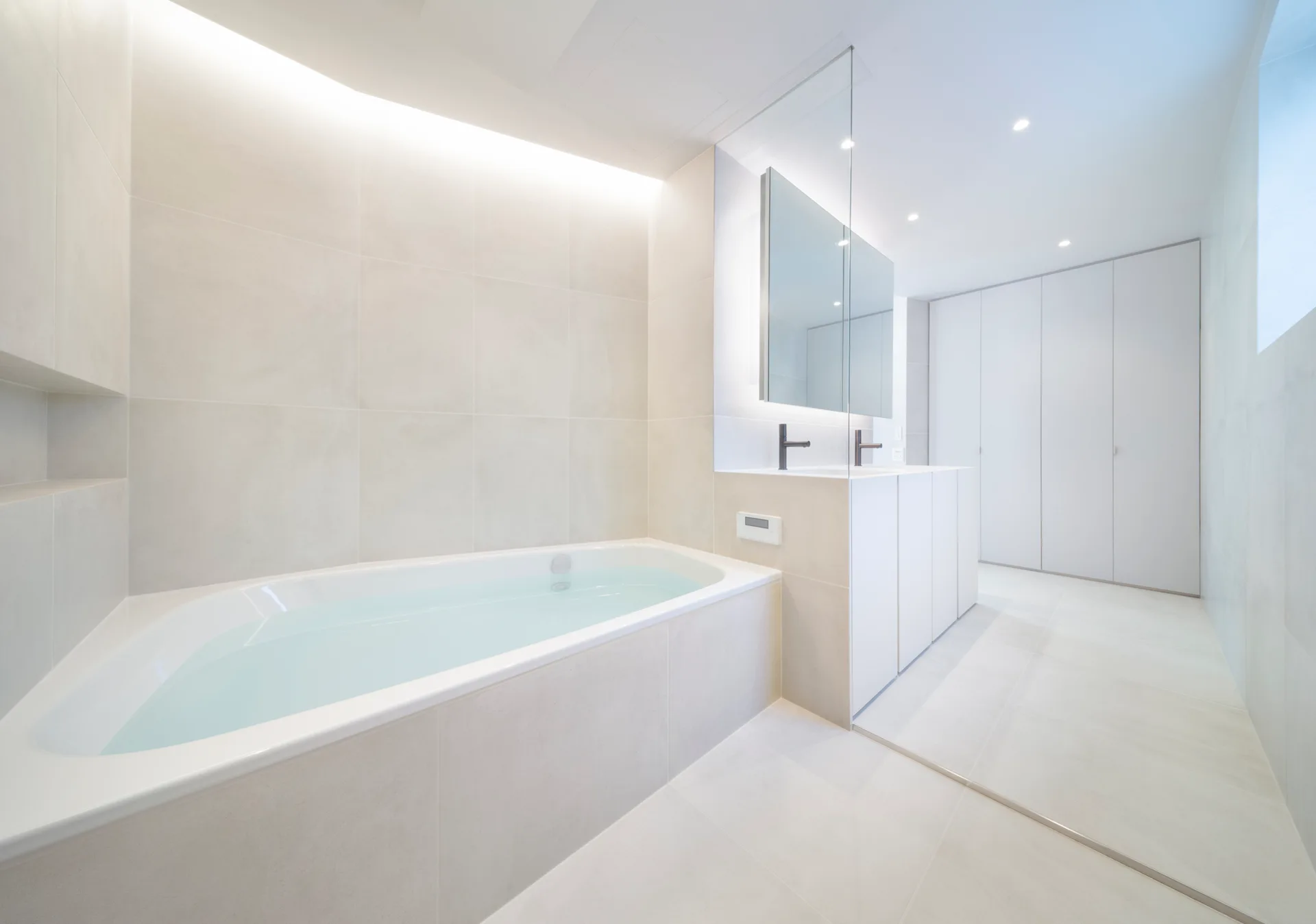 A minimalist bathroom features an integrated white bathtub, large-format beige tiles, a floating vanity with a backlit mirror, and dark faucets.