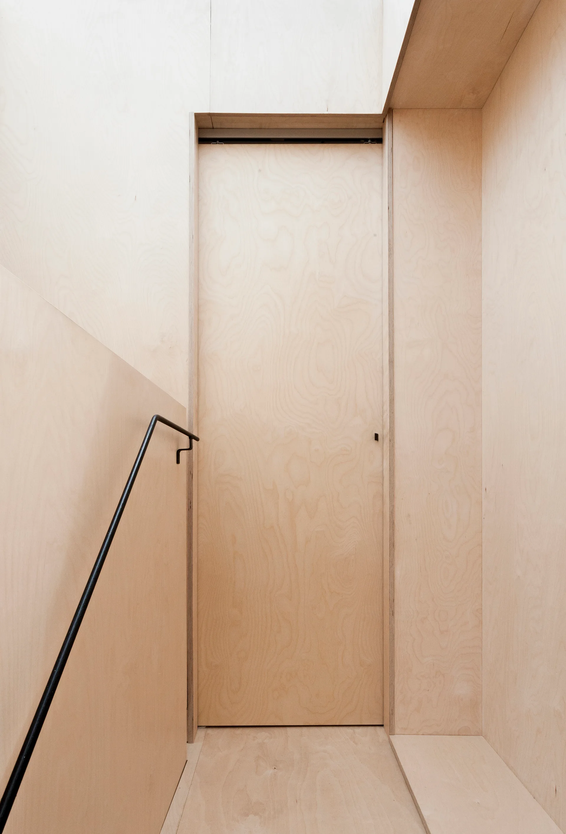A minimalist staircase landing clad in light plywood panels with a matching flush door and a thin black metal handrail.