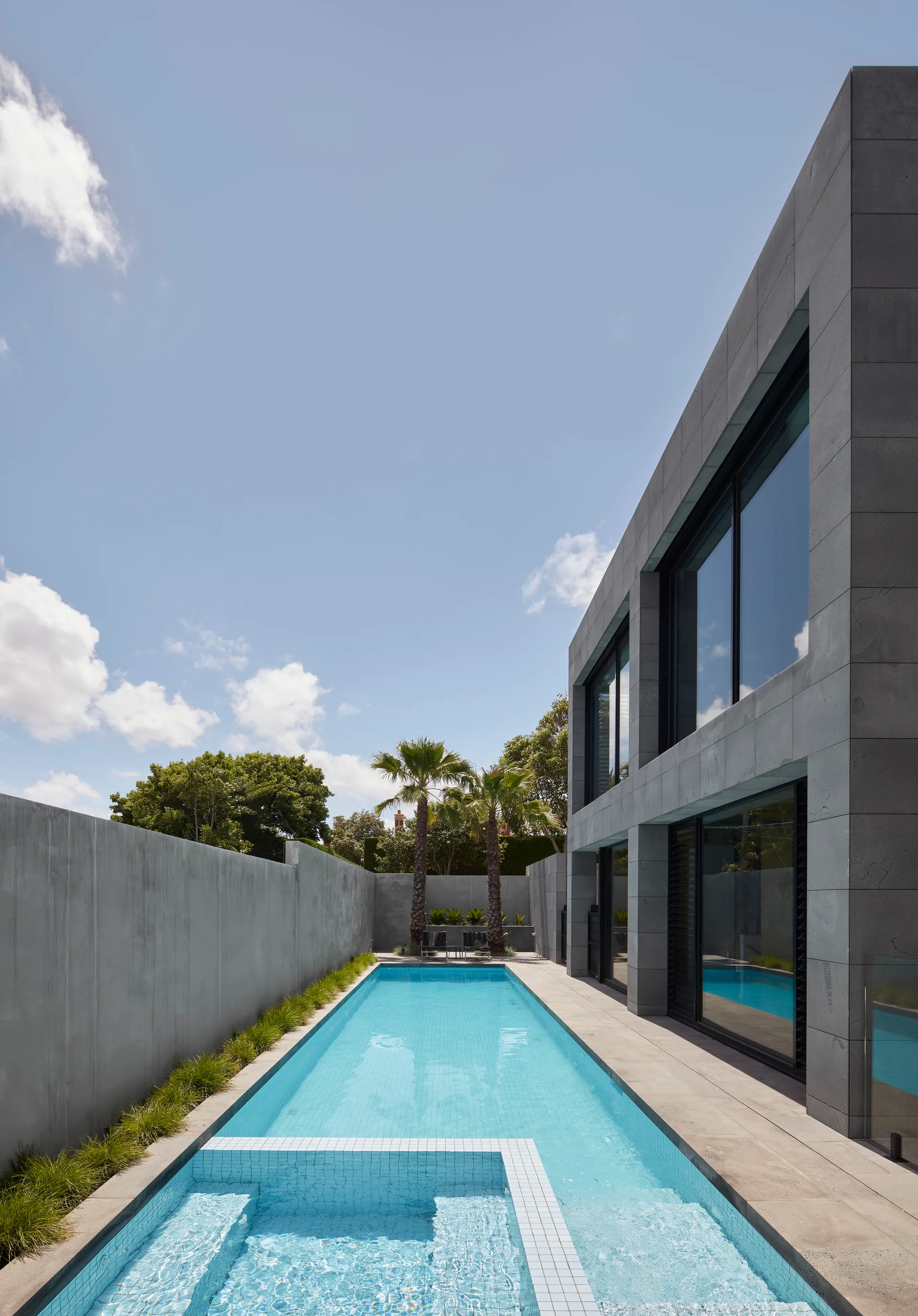 A narrow rectangular swimming pool runs between a tall concrete boundary wall and a modern grey stone building with large glass windows.