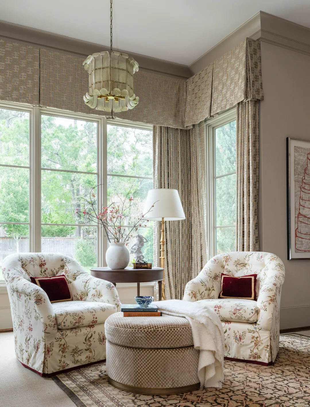 A traditional bedroom sitting area features two floral upholstered armchairs, a patterned ottoman, and structured window valances.