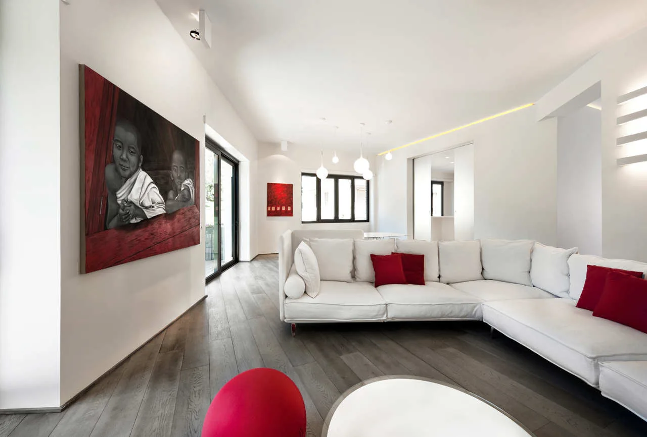 A minimalist white living room features a white L-shaped sectional sofa with red pillows on dark grey wood plank flooring near a large red painting.