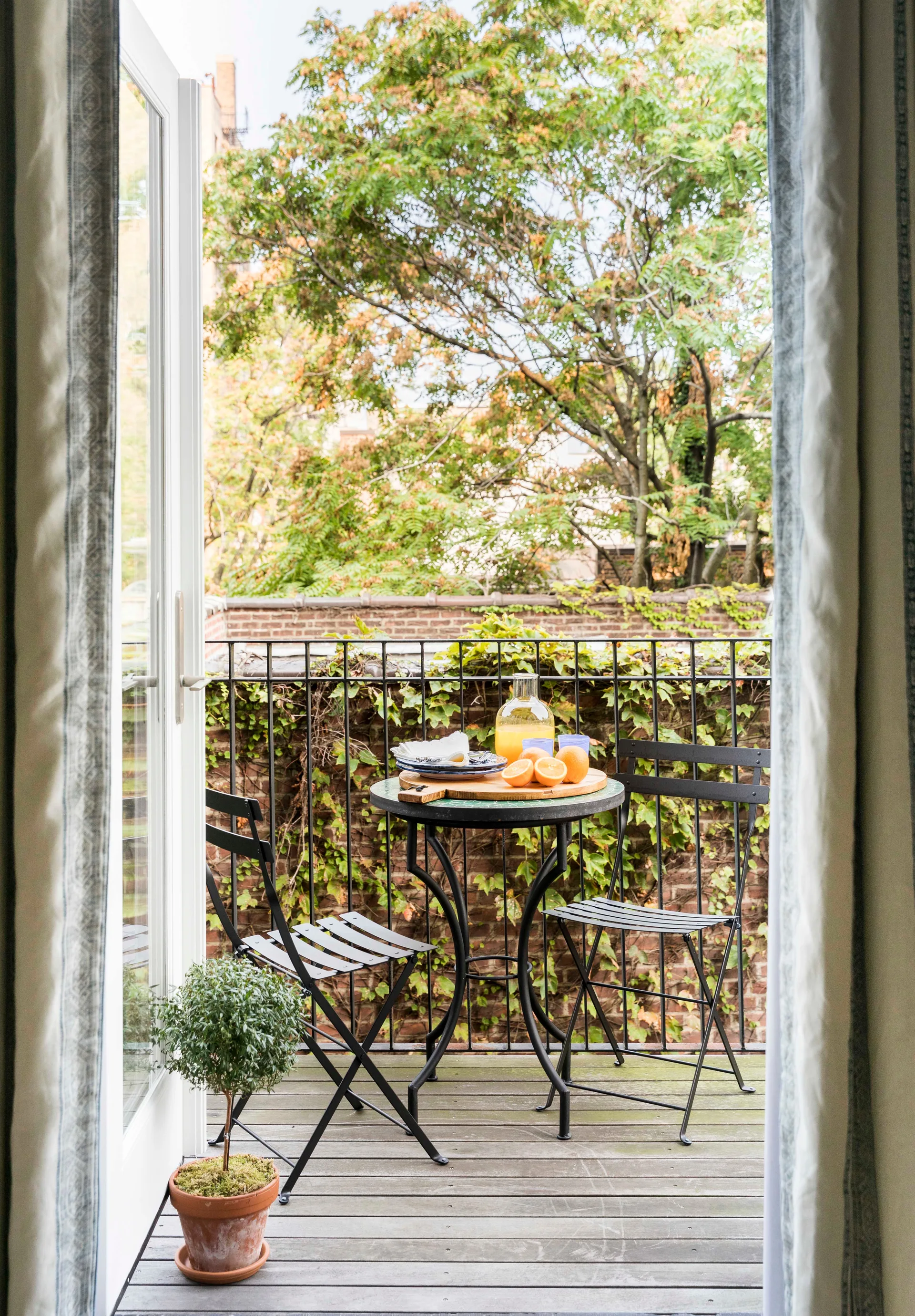 A view through open doors reveals a small balcony featuring a black metal bistro set, a small potted plant, and an ivy-covered brick wall.