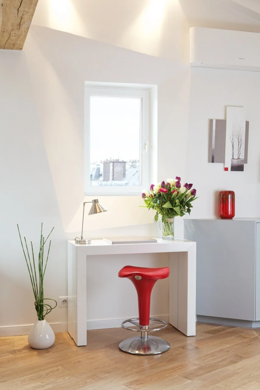 A small home office setup features a white console desk, a bright red saddle stool, and a desk lamp beneath a window under a sloped ceiling.