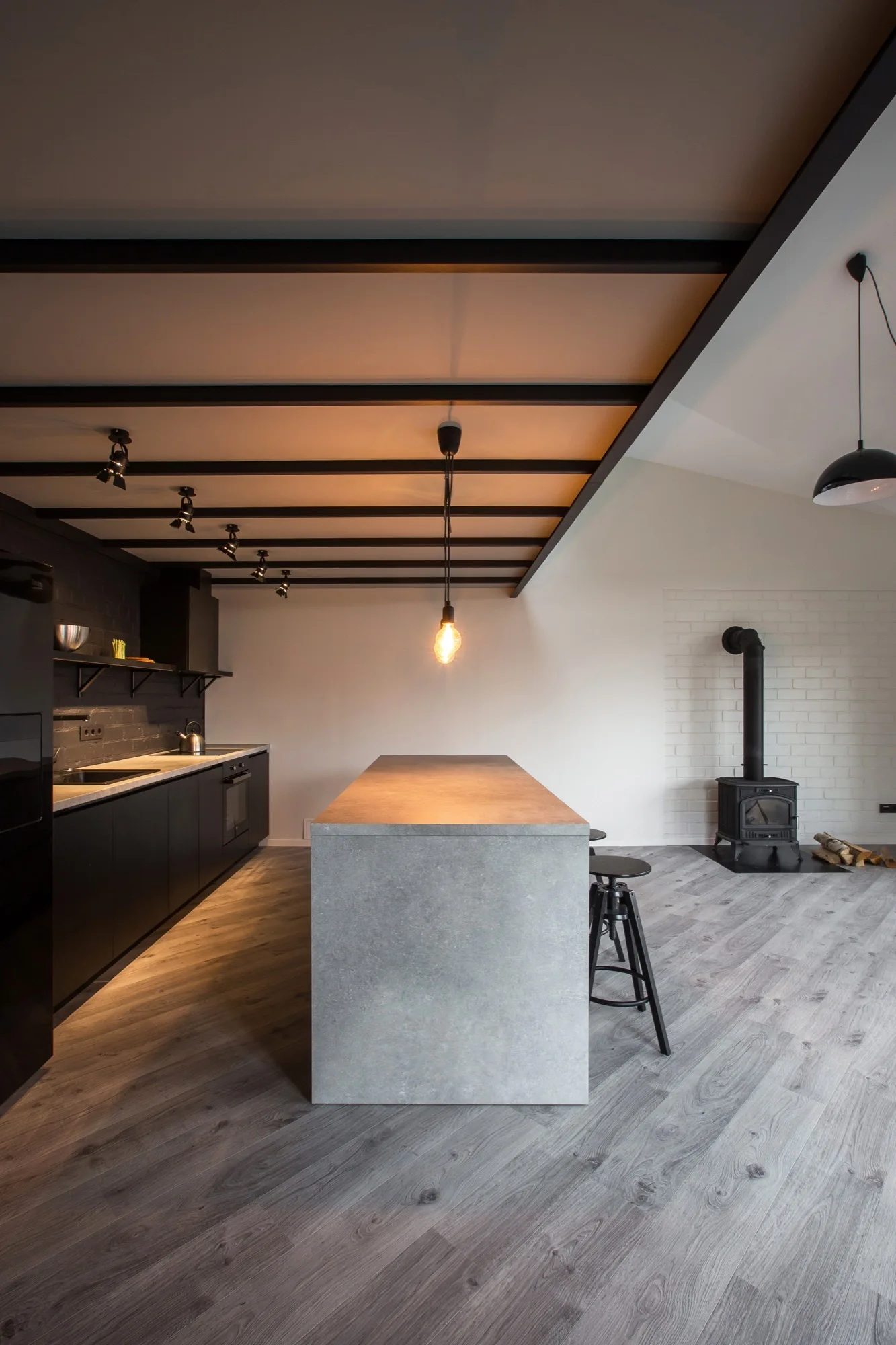 A modern kitchen features a grey stone-look island, black cabinetry, and black ceiling beams with a wood-burning stove in the background.