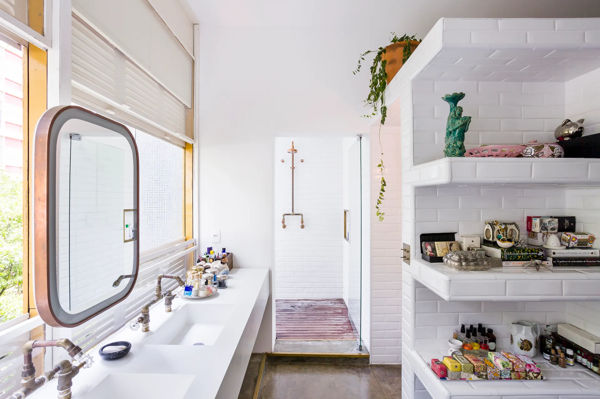 A white bathroom features a double-sink vanity with exposed copper plumbing, a glass shower, and open shelves wrapped in white subway tile.