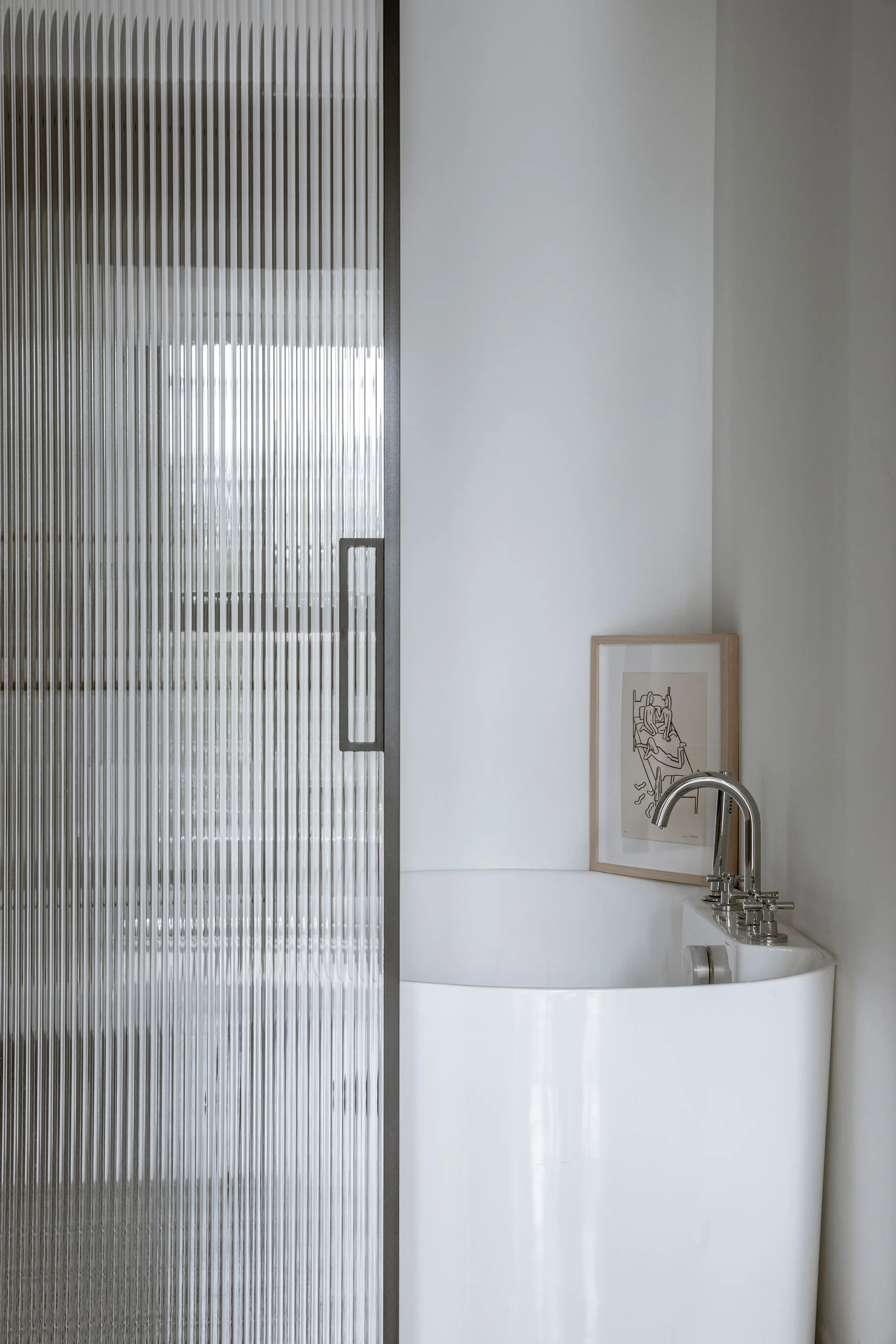A minimalist bathroom features a white soaking tub with a chrome faucet next to a textured fluted glass sliding door.