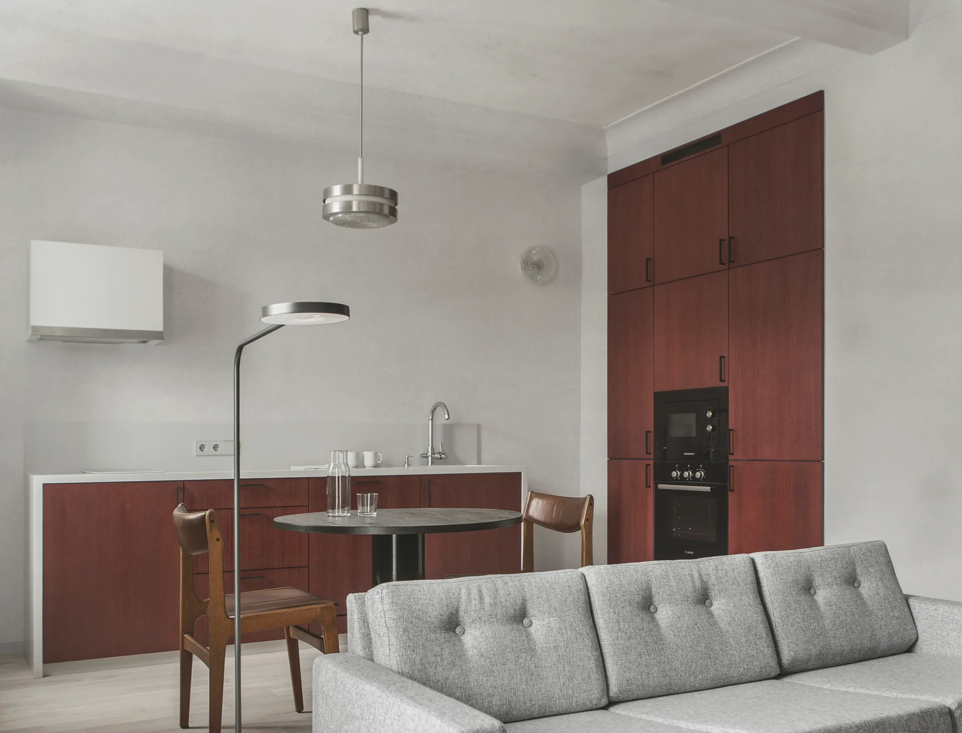 A minimalist studio apartment featuring deep reddish-brown kitchen cabinetry, a light grey tufted sofa, and a small round dining table.