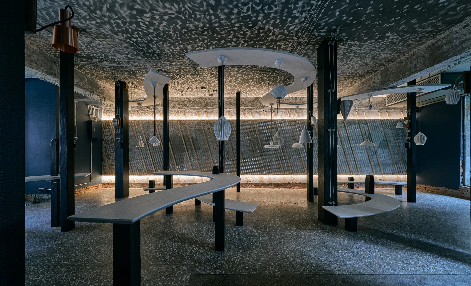 An industrial lounge space featuring curved grey benches mounted on black columns, ribbed pendant lights, and corrugated metal walls.