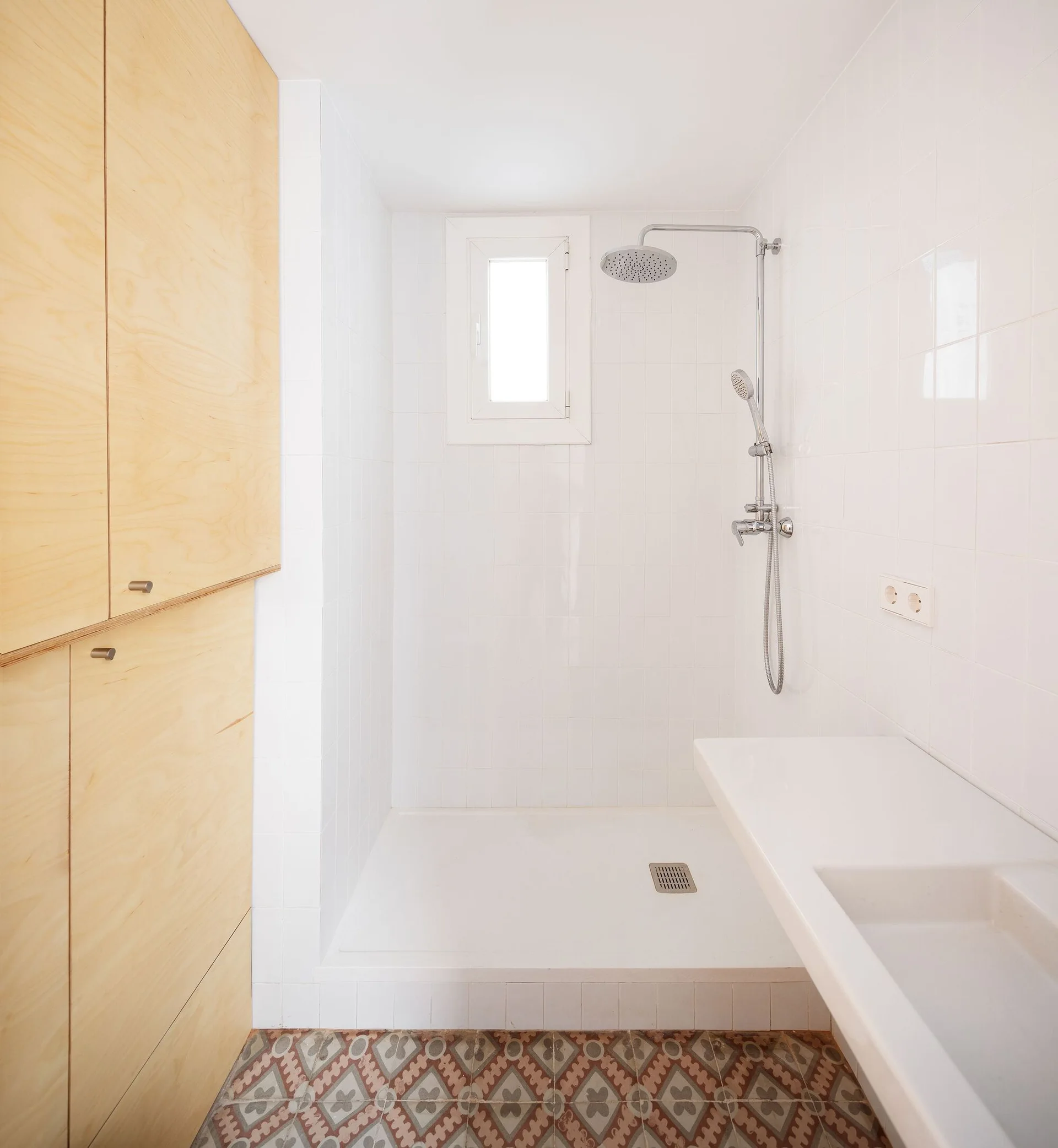 A modern white tiled bathroom features light wood cabinetry, a chrome shower fixture, and patterned geometric floor tiles.