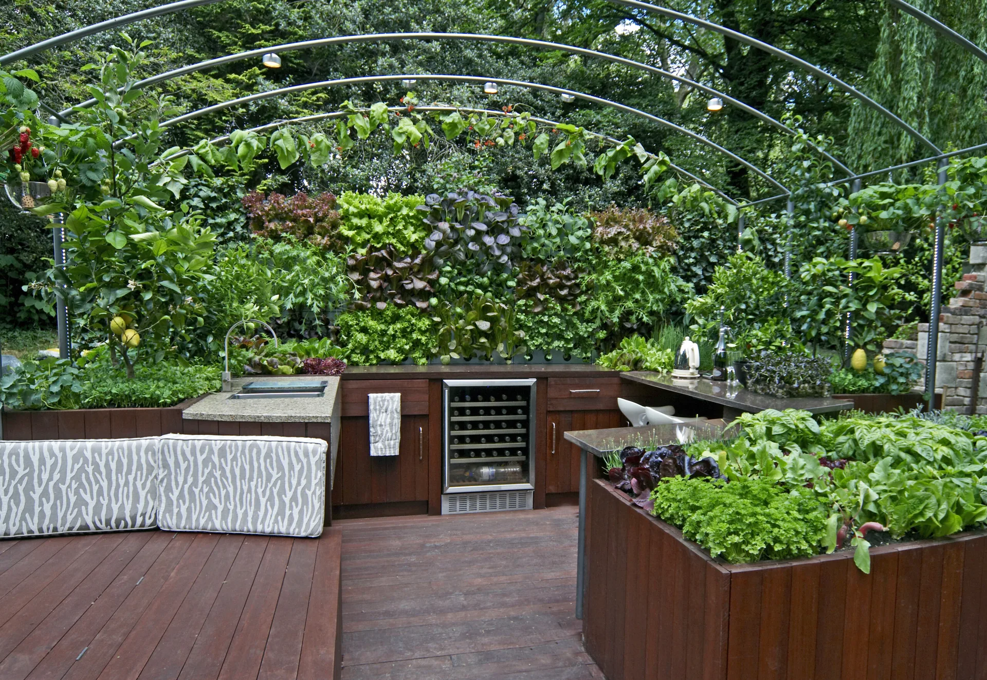 An outdoor kitchen featuring dark wood cabinetry, a built-in wine cooler, and a lush vertical vegetable garden under an arched metal trellis.