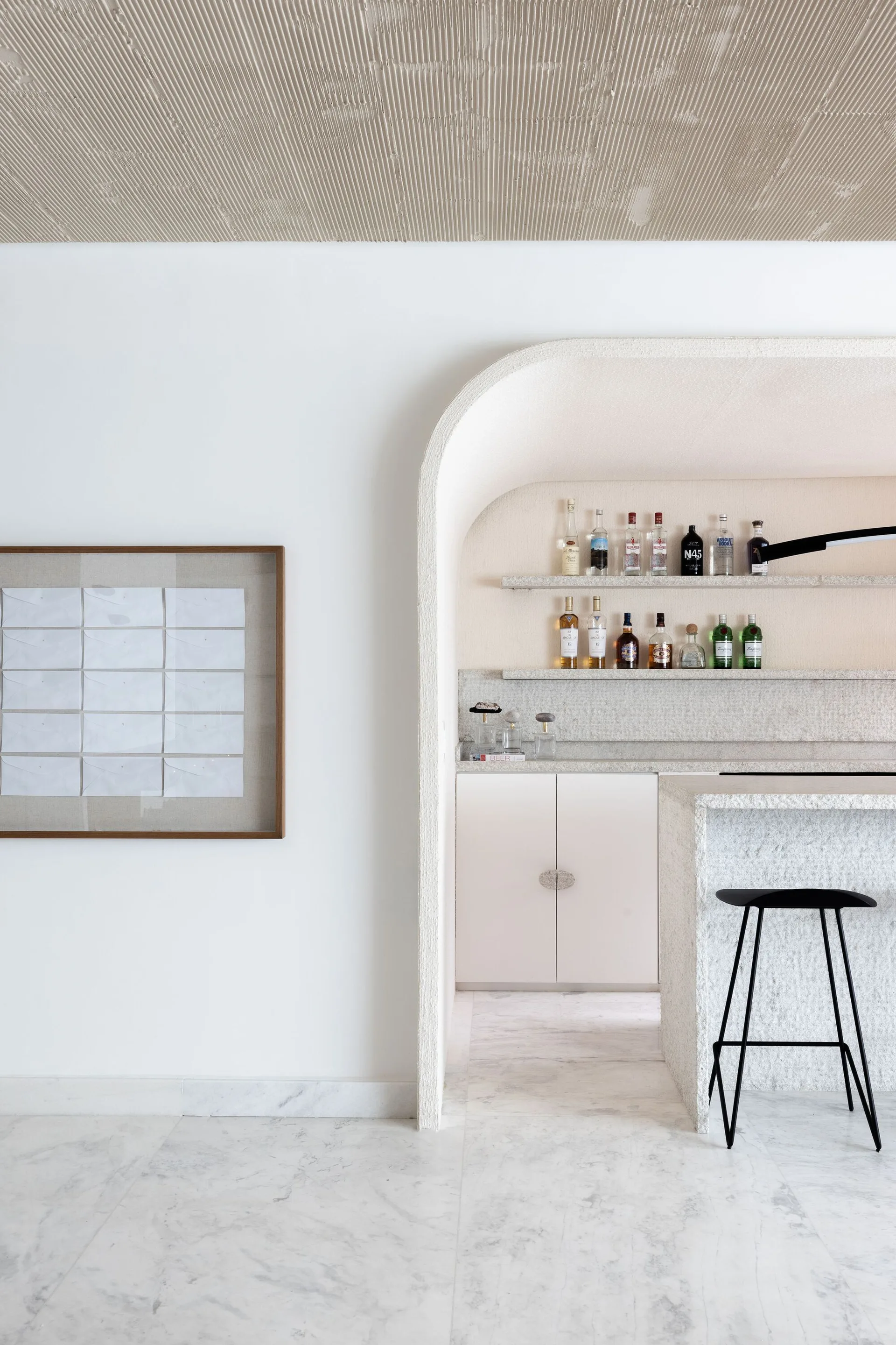 A minimalist home bar framed by a rough-textured stone archway featuring floating shelves with bottles and a black bar stool.