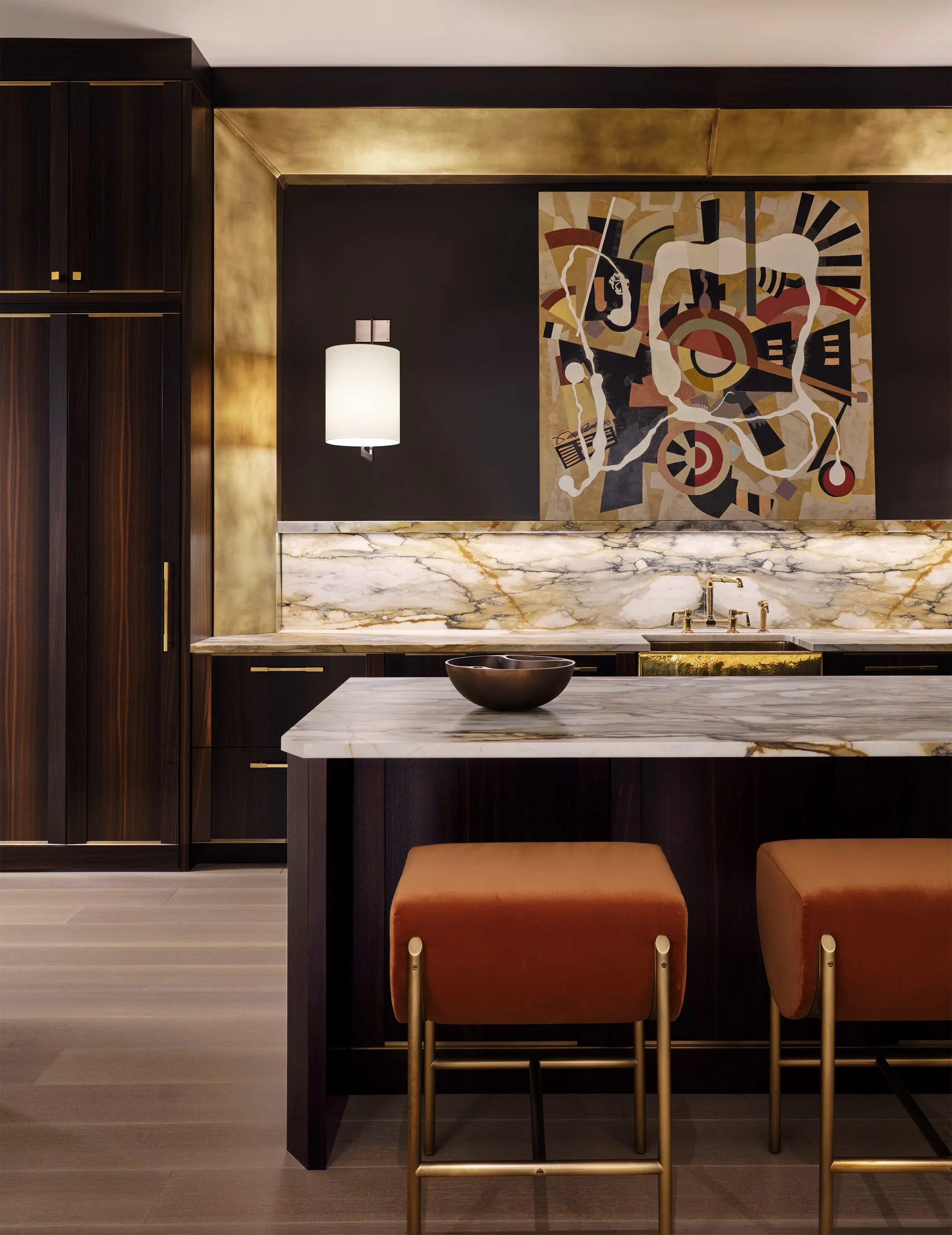 A dark wood home bar with a marble countertop, orange velvet stools, and a gold-lined niche holding an abstract painting.