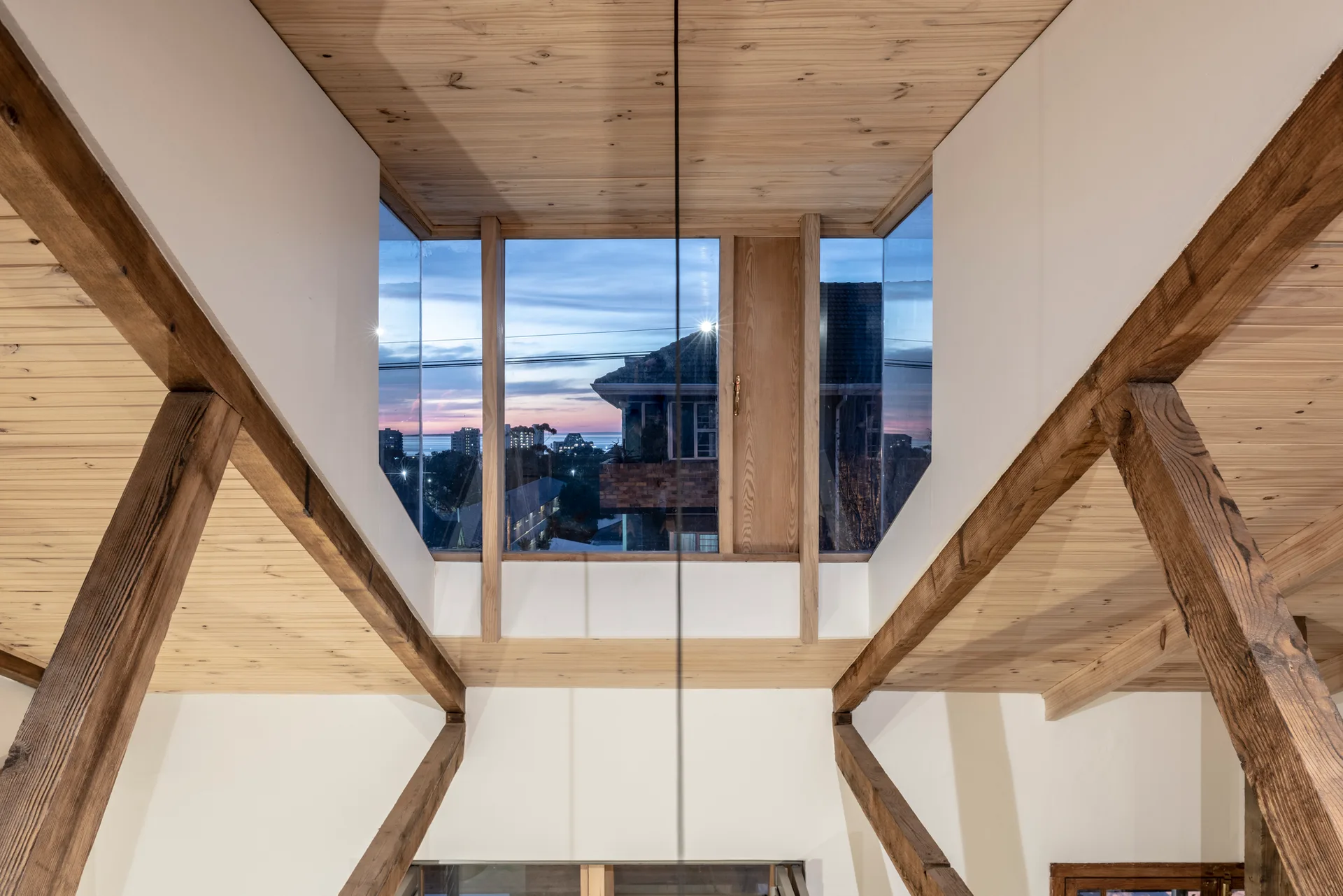 A residential attic interior features a vaulted wood plank ceiling with thick exposed beams framing a large window that overlooks a city skyline at twilight.