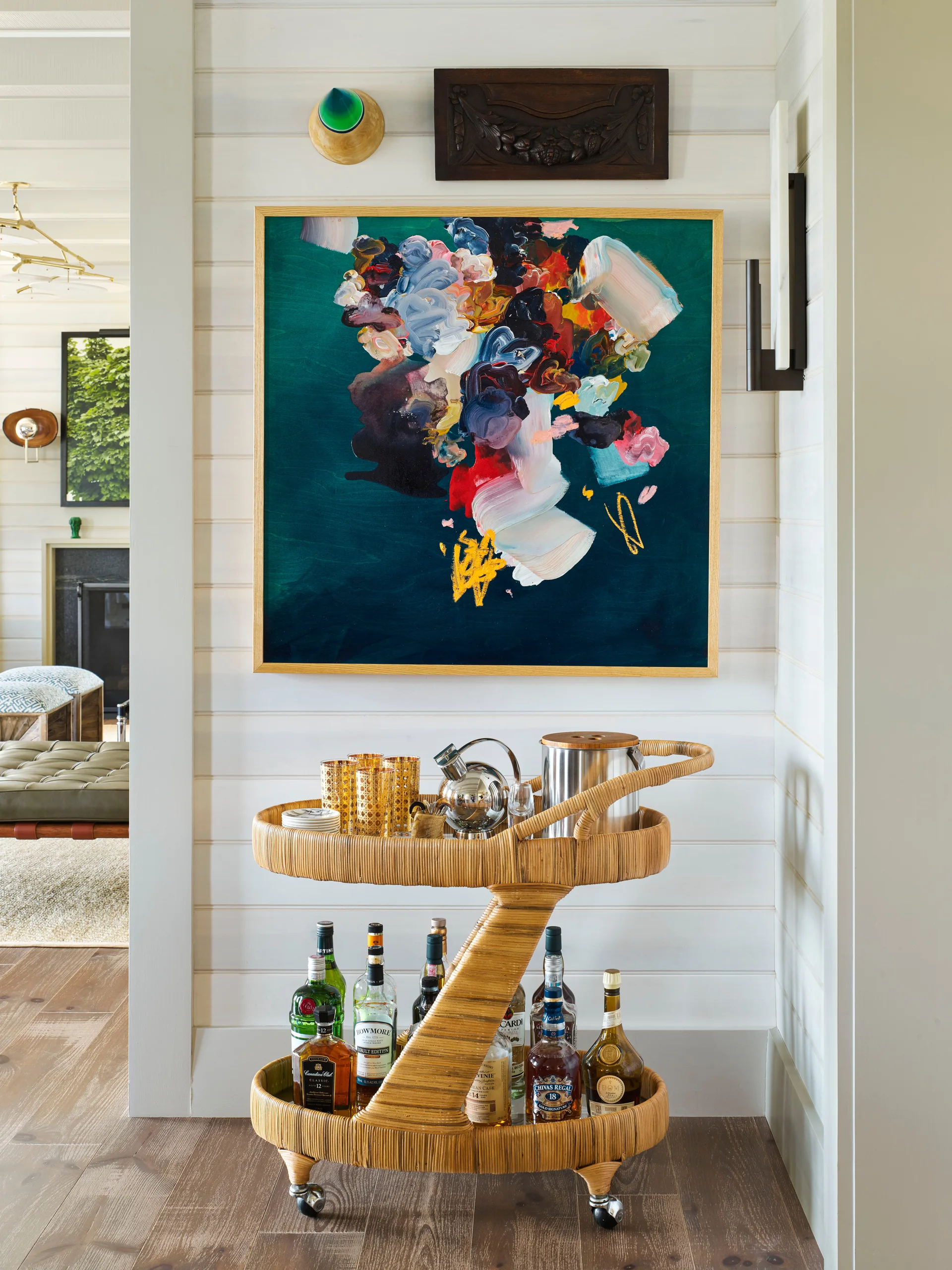 A two-tier rattan bar cart stocked with bottles stands against a white shiplap wall beneath a colorful abstract painting.