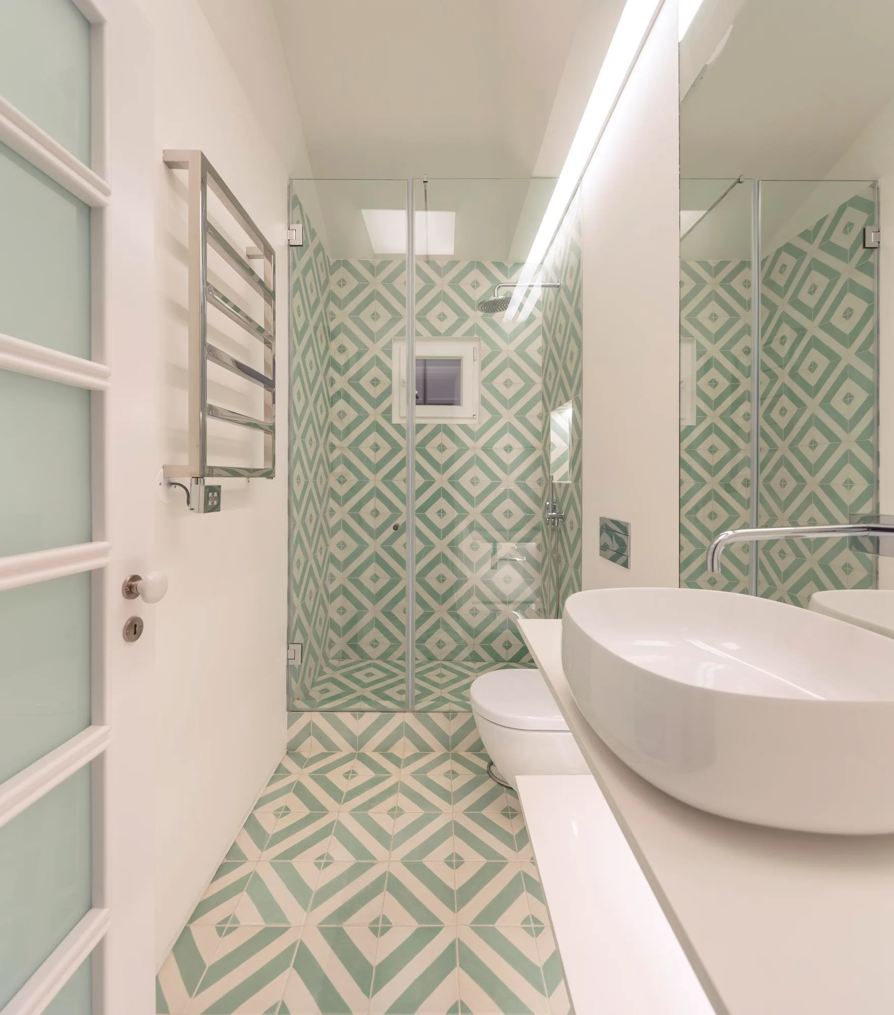 A narrow bathroom features mint green and white geometric floor tiles that continue into a glass shower, with a white vessel sink on the vanity.