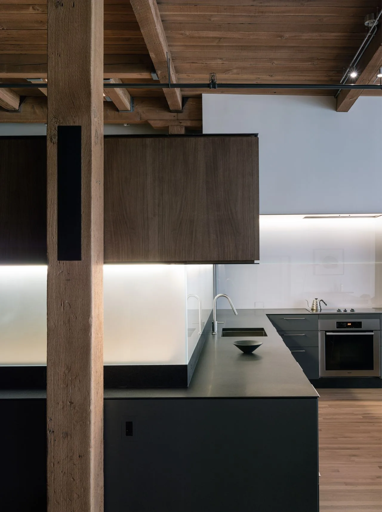 A modern kitchen features dark charcoal cabinetry, an illuminated frosted glass backsplash, and an exposed heavy timber ceiling with wooden beams.