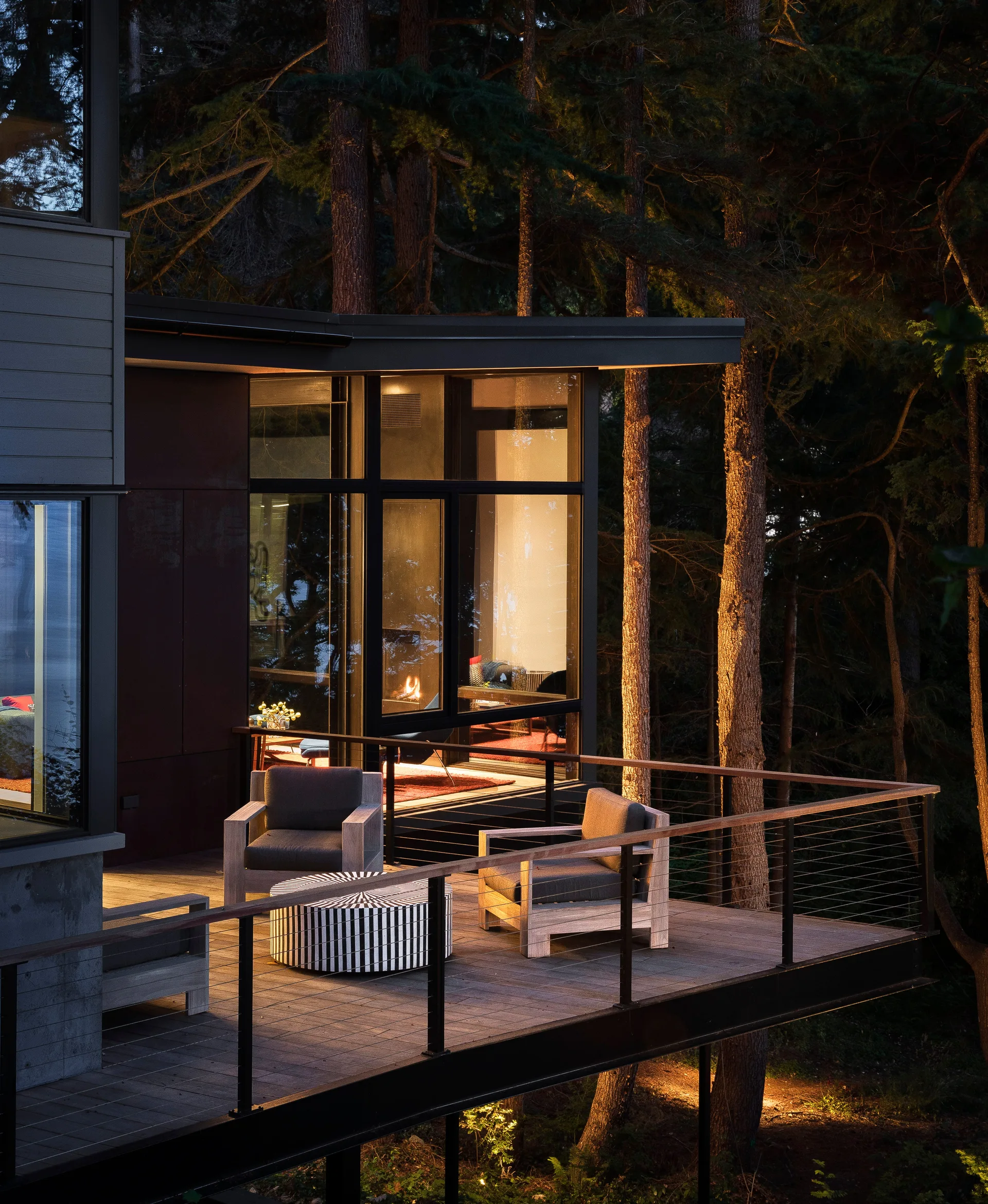 A raised wooden terrace features two gray armchairs and a striped coffee table, surrounded by forest trees with warm light glowing from glass walls.