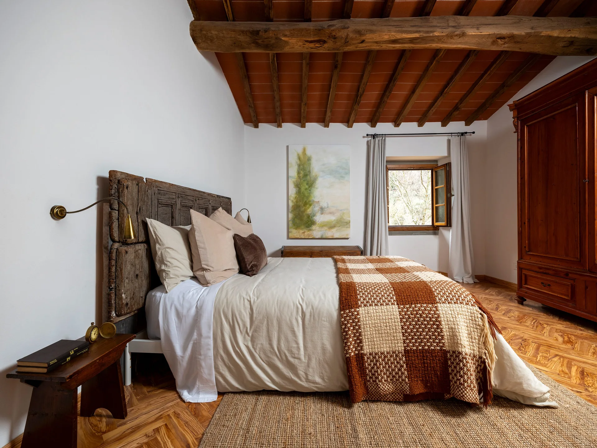 A rustic bedroom features a bed with a textured wood headboard, a checkered blanket, exposed ceiling beams, and herringbone wood floors.