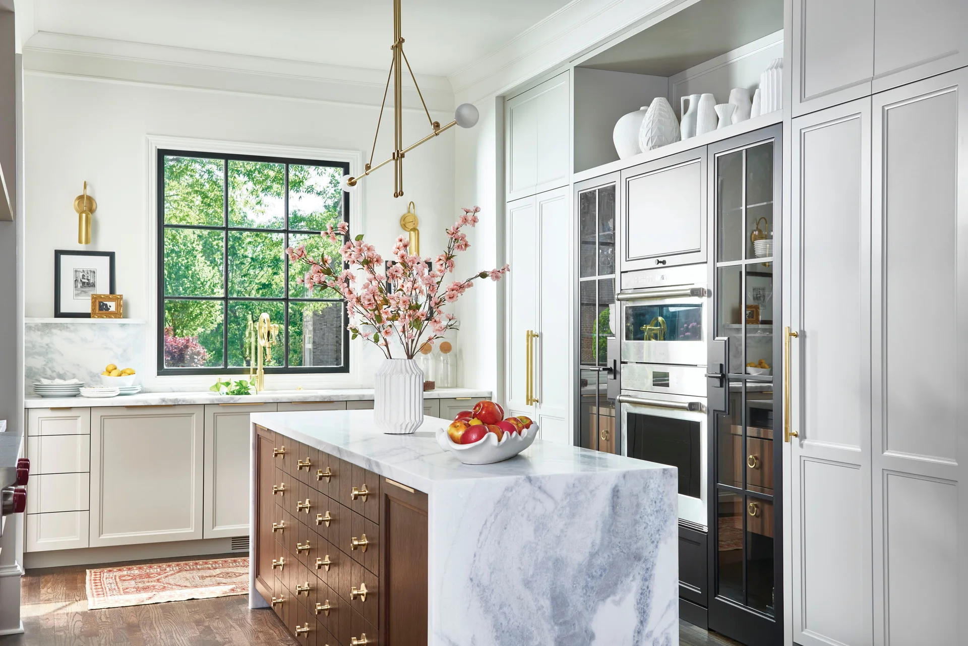 A bright kitchen features a marble-topped wood island with numerous small brass-handled drawers, white cabinetry, and built-in double ovens.