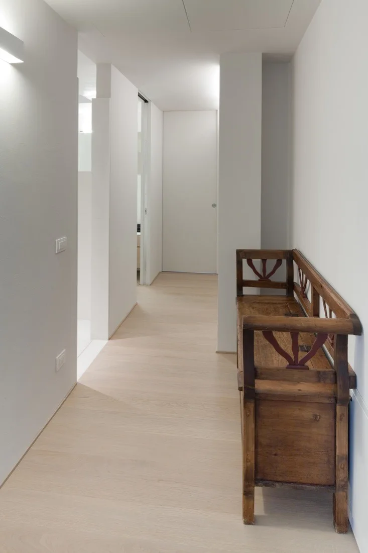 A bright white minimalist hallway features light wood floors and an antique-style dark wood bench positioned against the wall.