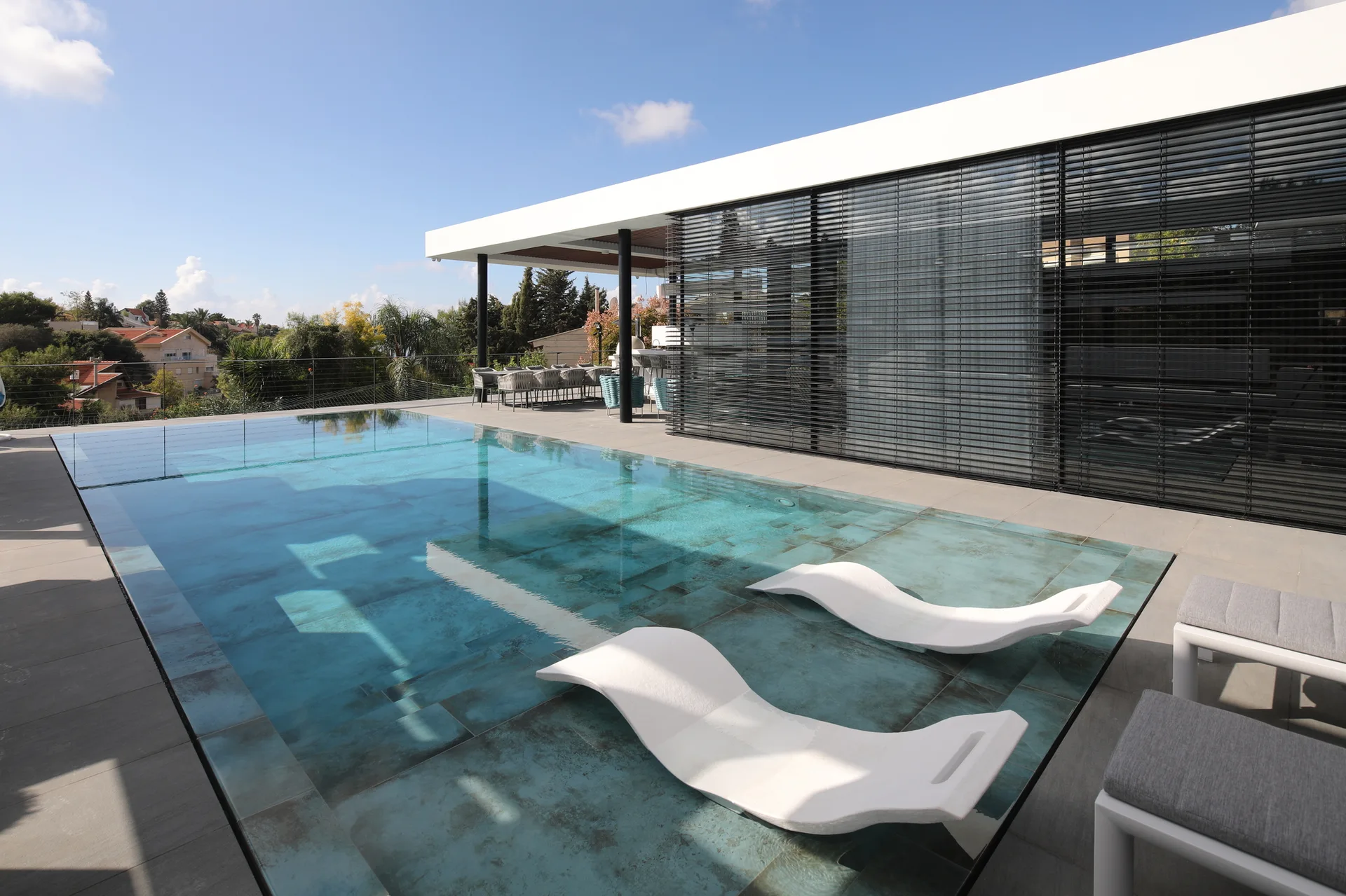 A modern outdoor terrace features a tiled swimming pool with two submerged white lounge chairs alongside a grey stone patio and cantilevered roof.