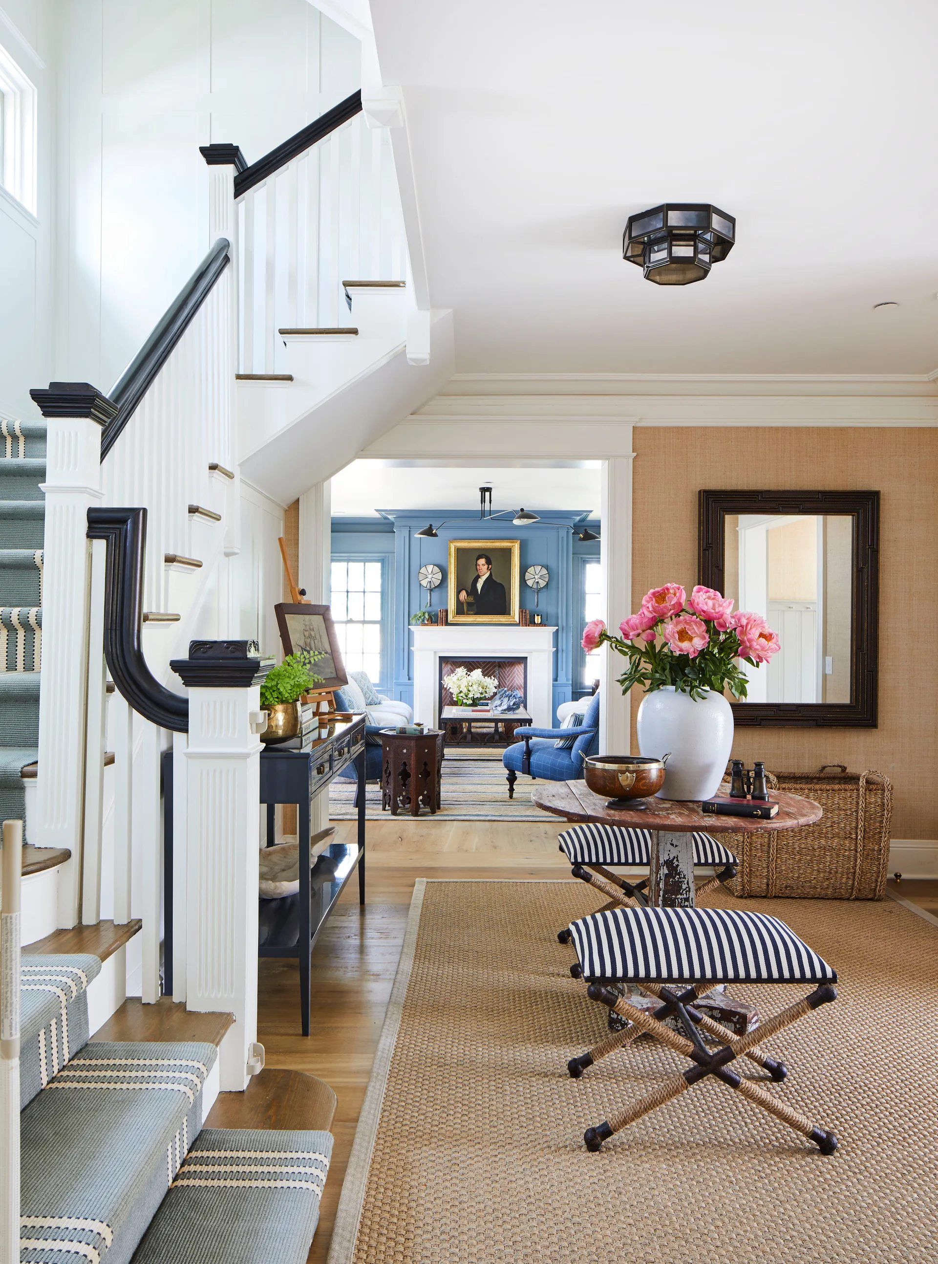 A bright foyer features a staircase with a striped runner, a small round table with pink flowers, and a glimpse of a blue paneled living room.