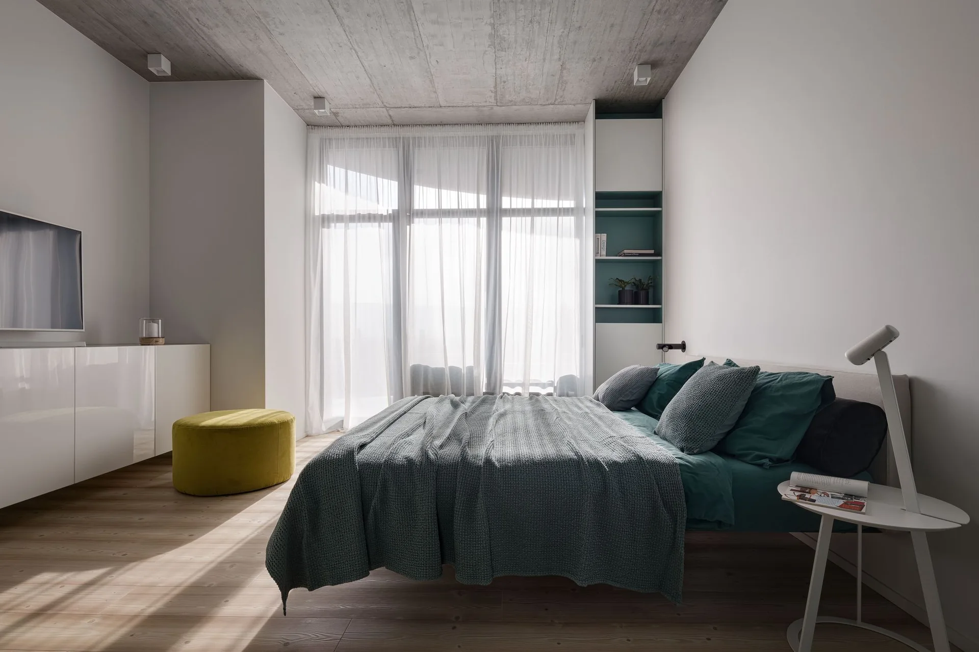 A modern bedroom features a low bed with teal bedding, an exposed concrete ceiling, sheer white curtains, and a round yellow ottoman.