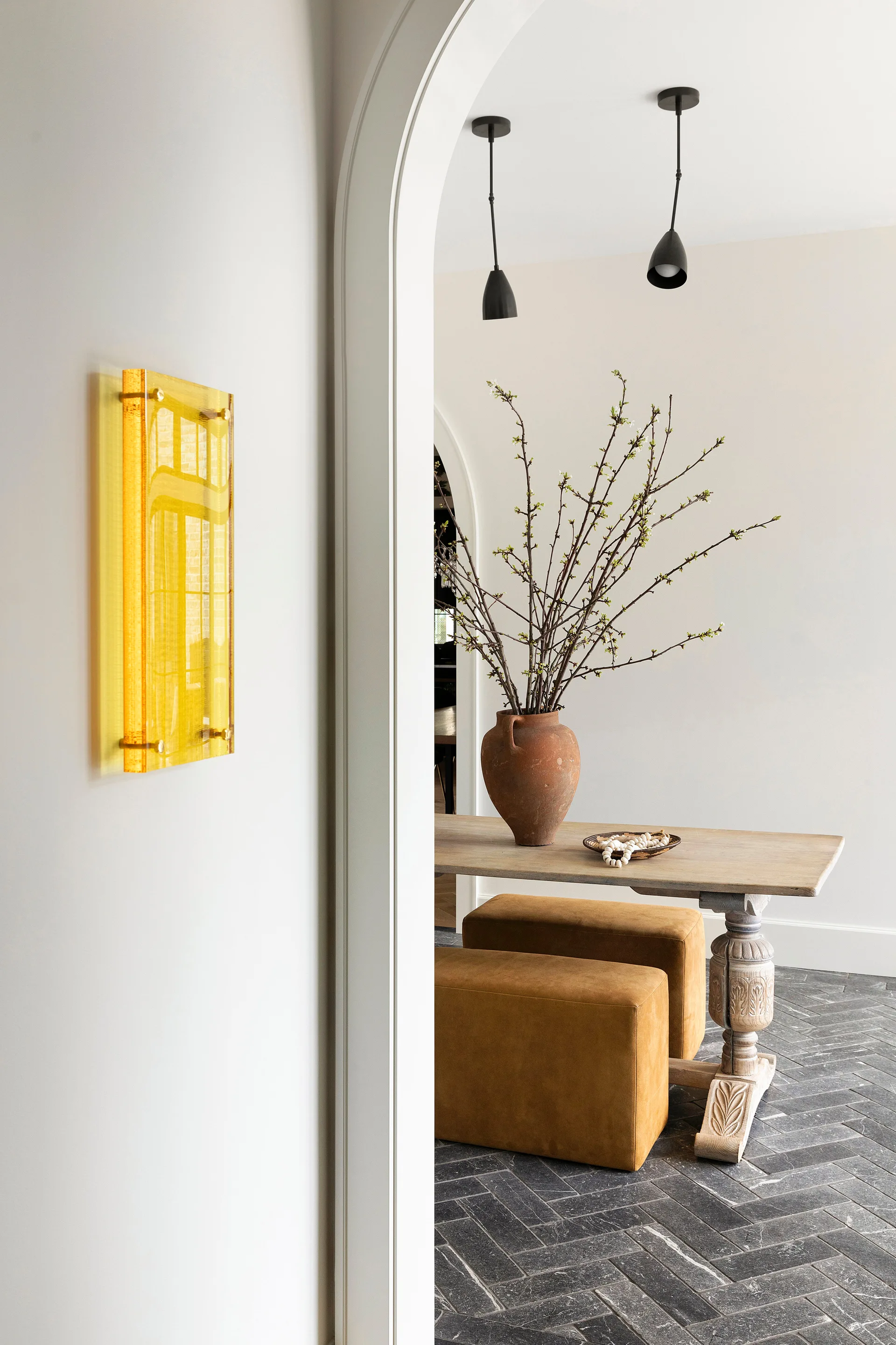 An arched doorway looks into an entryway featuring a wooden console table with ochre ottomans beneath, a terracotta vase, and a herringbone stone floor.