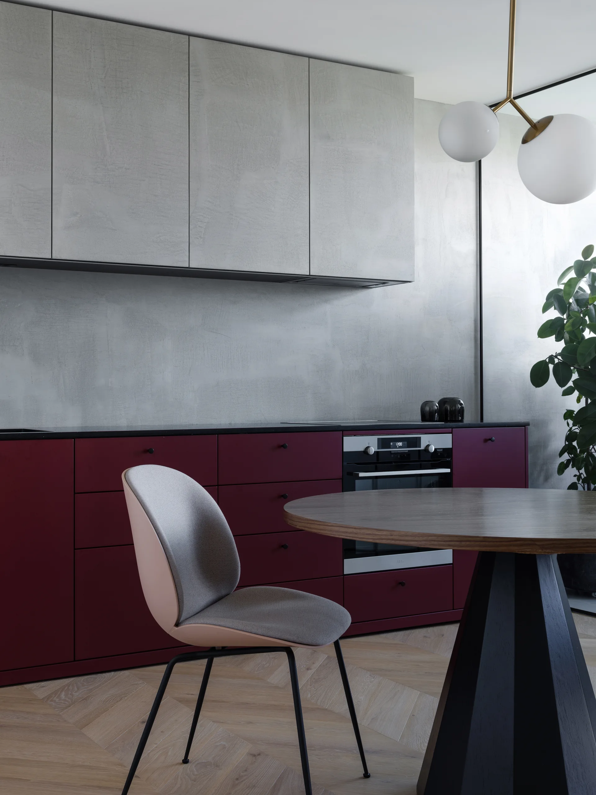 A minimalist kitchen features burgundy lower cabinets, textured grey upper cabinets, a round wood table, and herringbone pattern wood flooring.