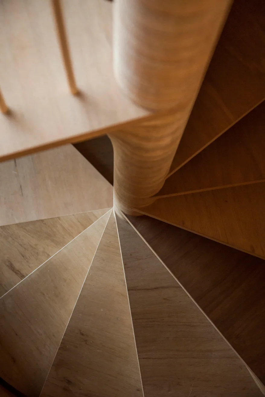 Overhead view of a minimalist spiral staircase crafted from light-toned plywood with a central column and fan-shaped steps.