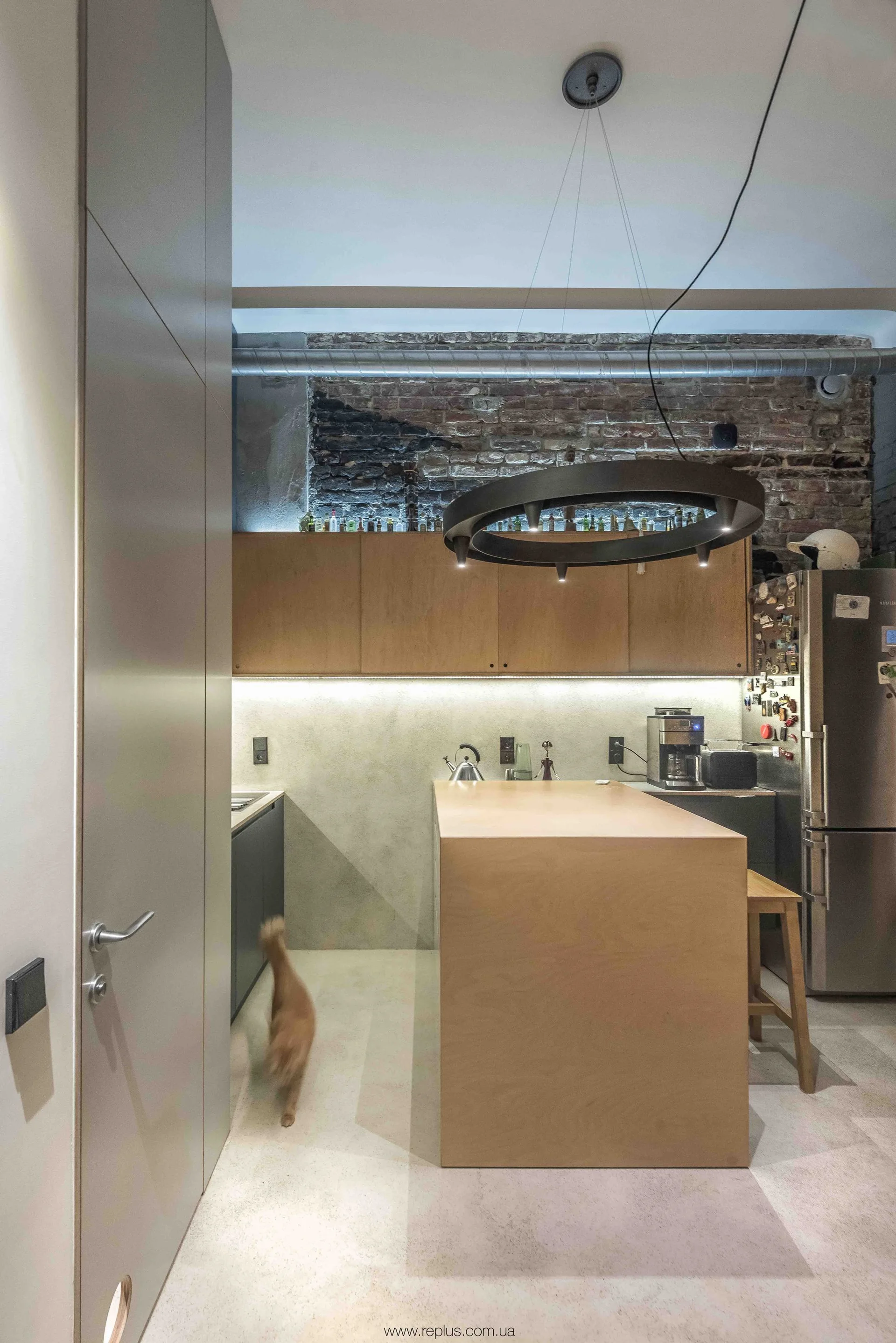 A modern industrial kitchen features a wooden island, matching upper cabinets under an exposed brick wall, and a large circular pendant light.