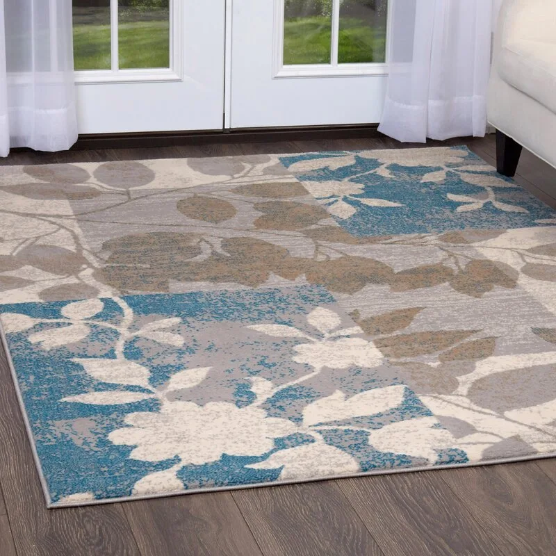 A rectangular area rug with a blue and grey botanical patchwork pattern rests on a dark hardwood floor in front of white glass doors.