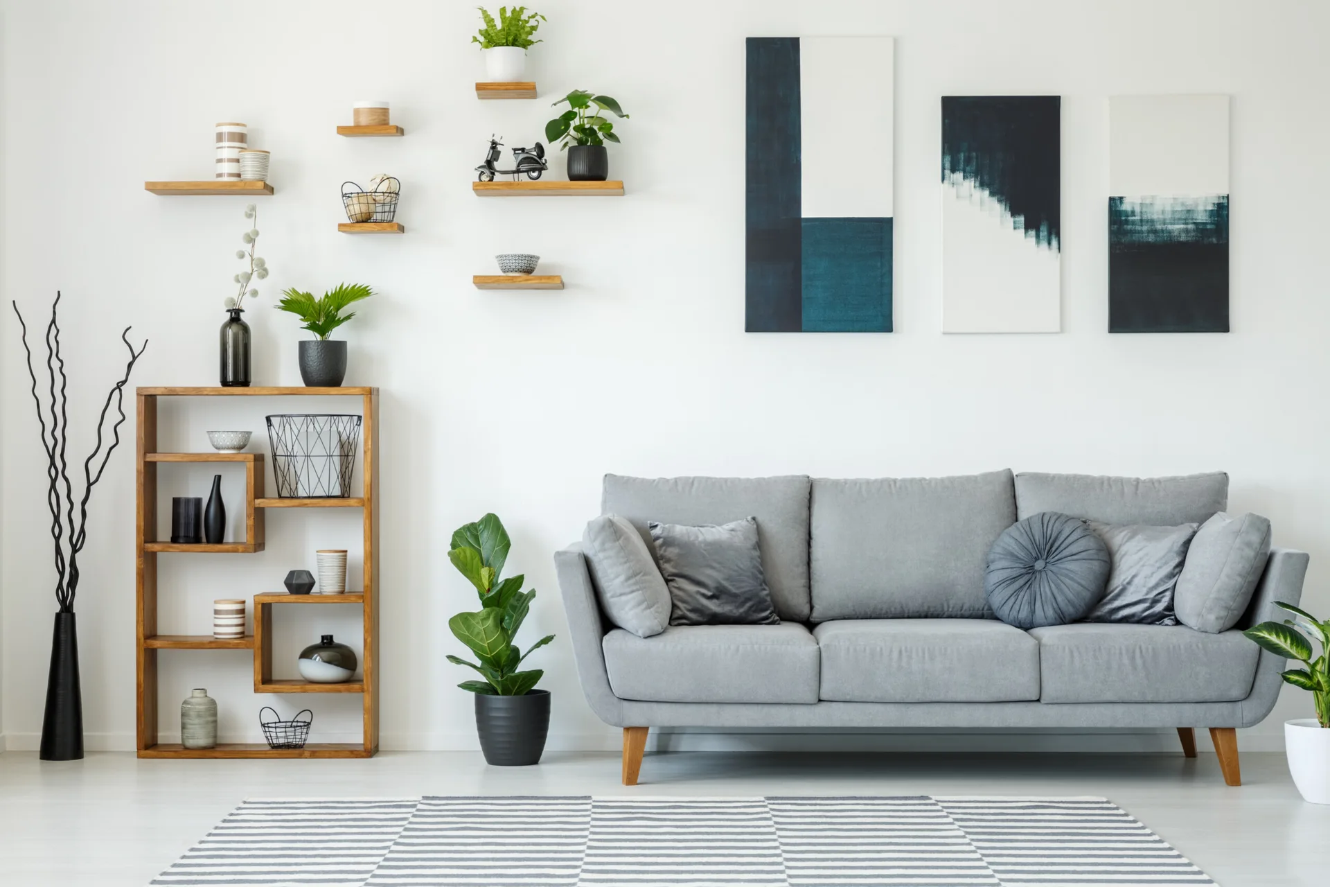 A minimalist living room features a gray fabric sofa against a white wall decorated with three vertical canvases and staggered wooden floating shelves.