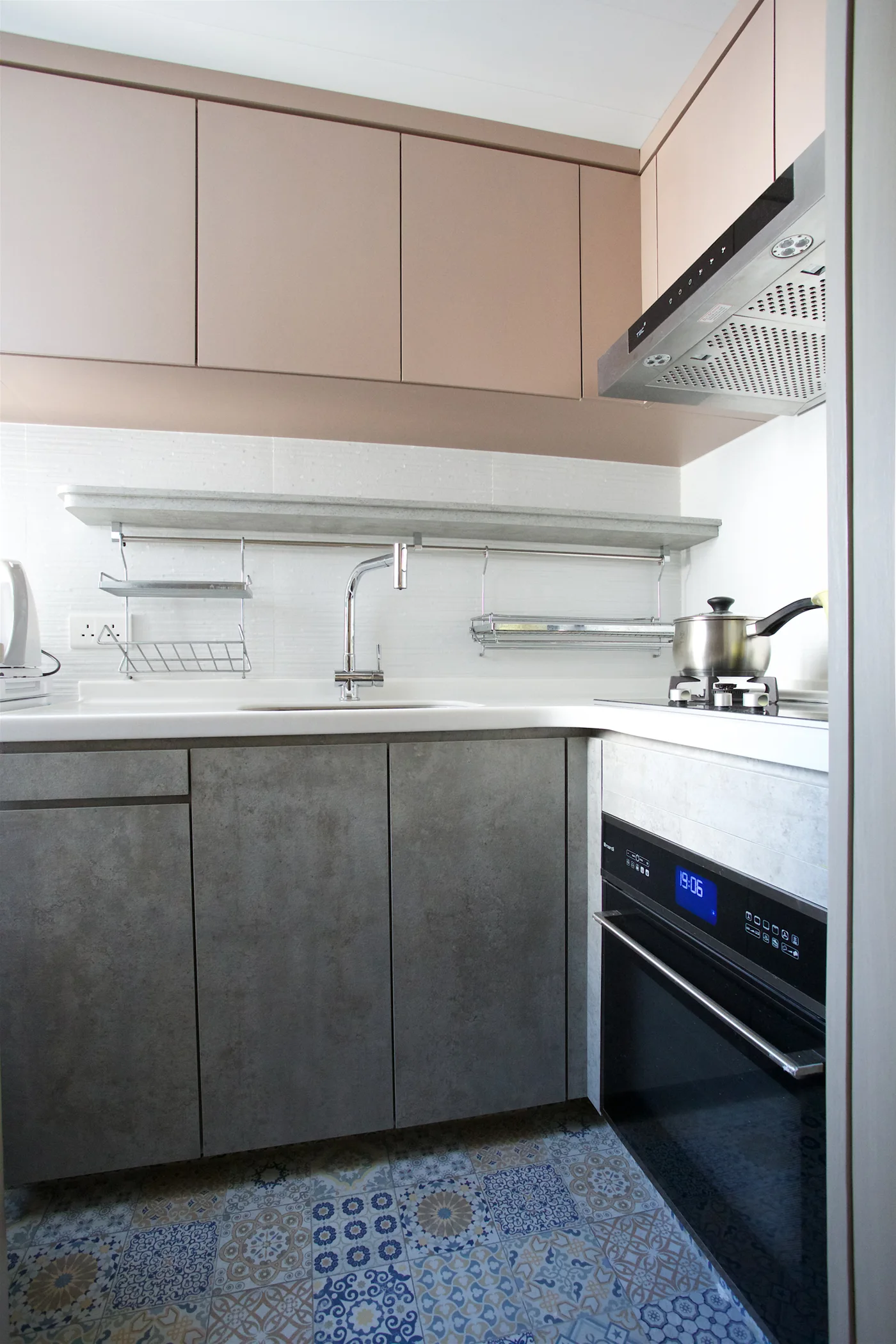 A compact kitchen features blush pink upper cabinets, dark grey textured lower cabinets, a white tiled backsplash, and patterned blue ceramic floor tiles.