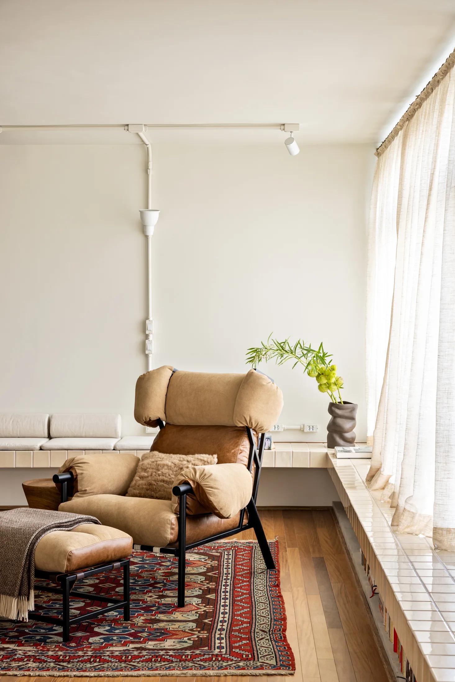 A tan and brown leather armchair with an ottoman sits on a patterned red rug next to a low tiled bench under sheer curtains.