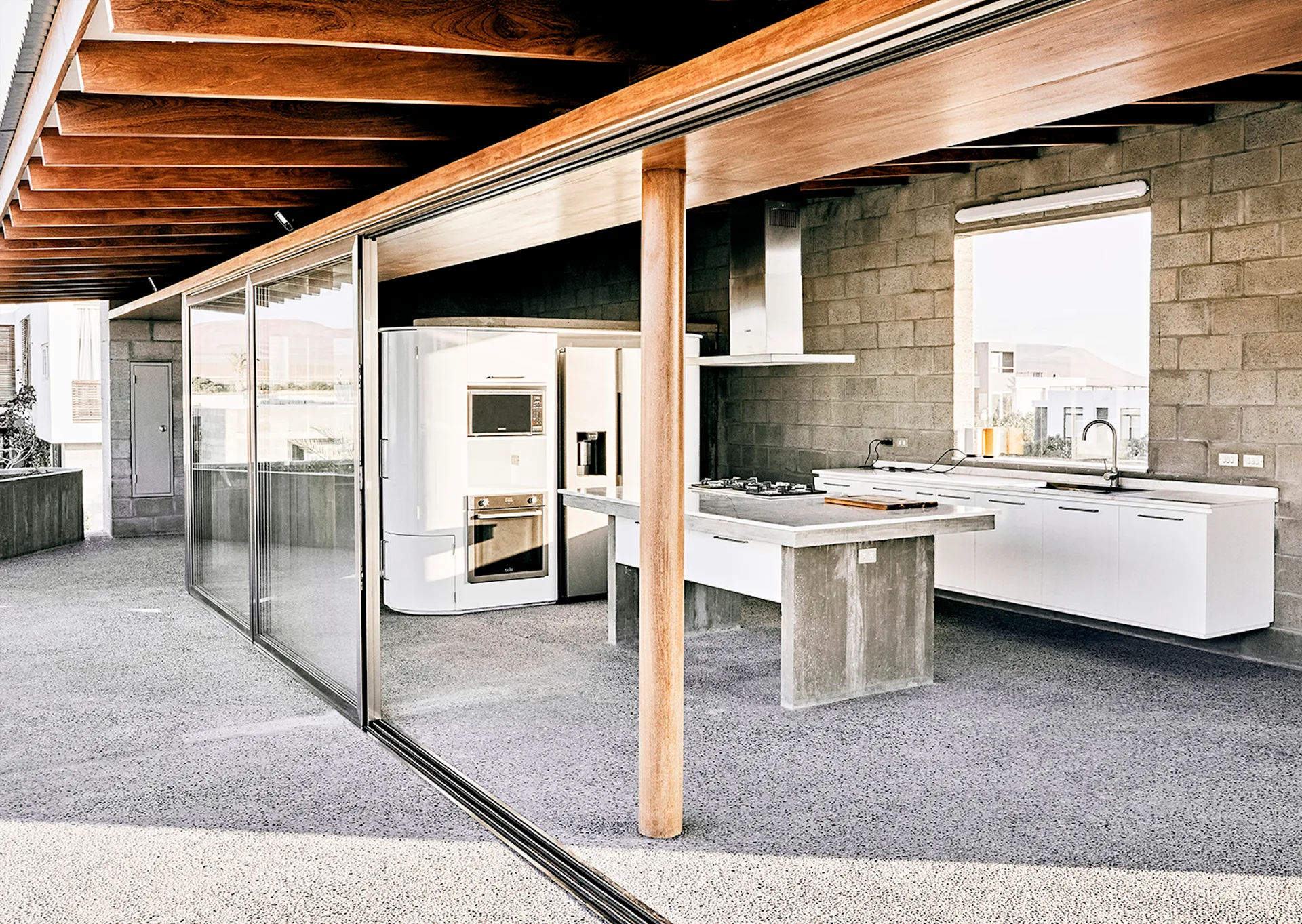 An open-concept kitchen features a concrete-topped island, white cabinets, and an exposed masonry block wall beneath a timber-raftered ceiling.