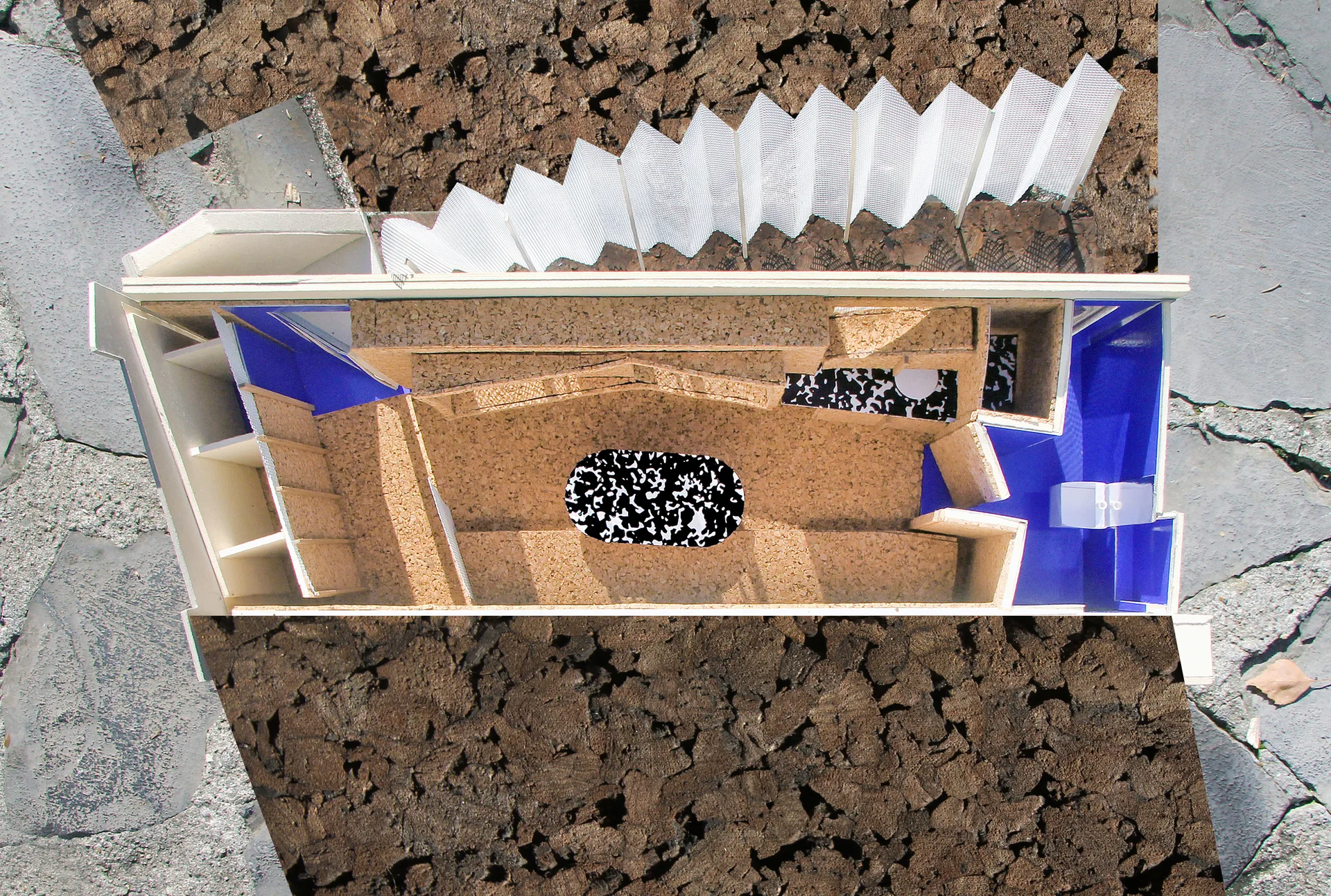 An overhead view of an architectural scale model featuring cork floors, blue partition walls, a white pleated screen, and a speckled rug.