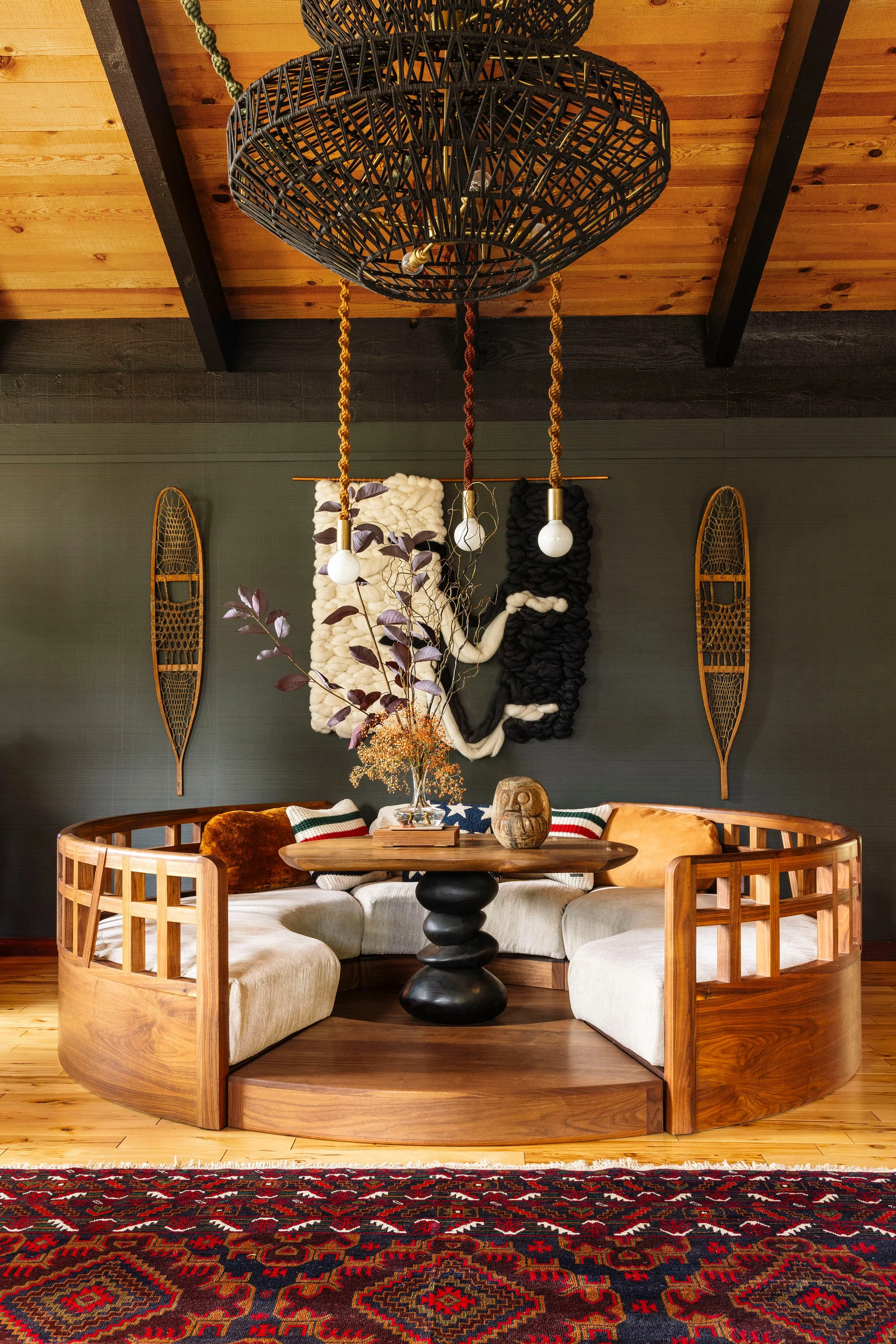 A circular wooden conversation pit with cushions sits in a cabin living room featuring dark green walls and a planked wood ceiling.