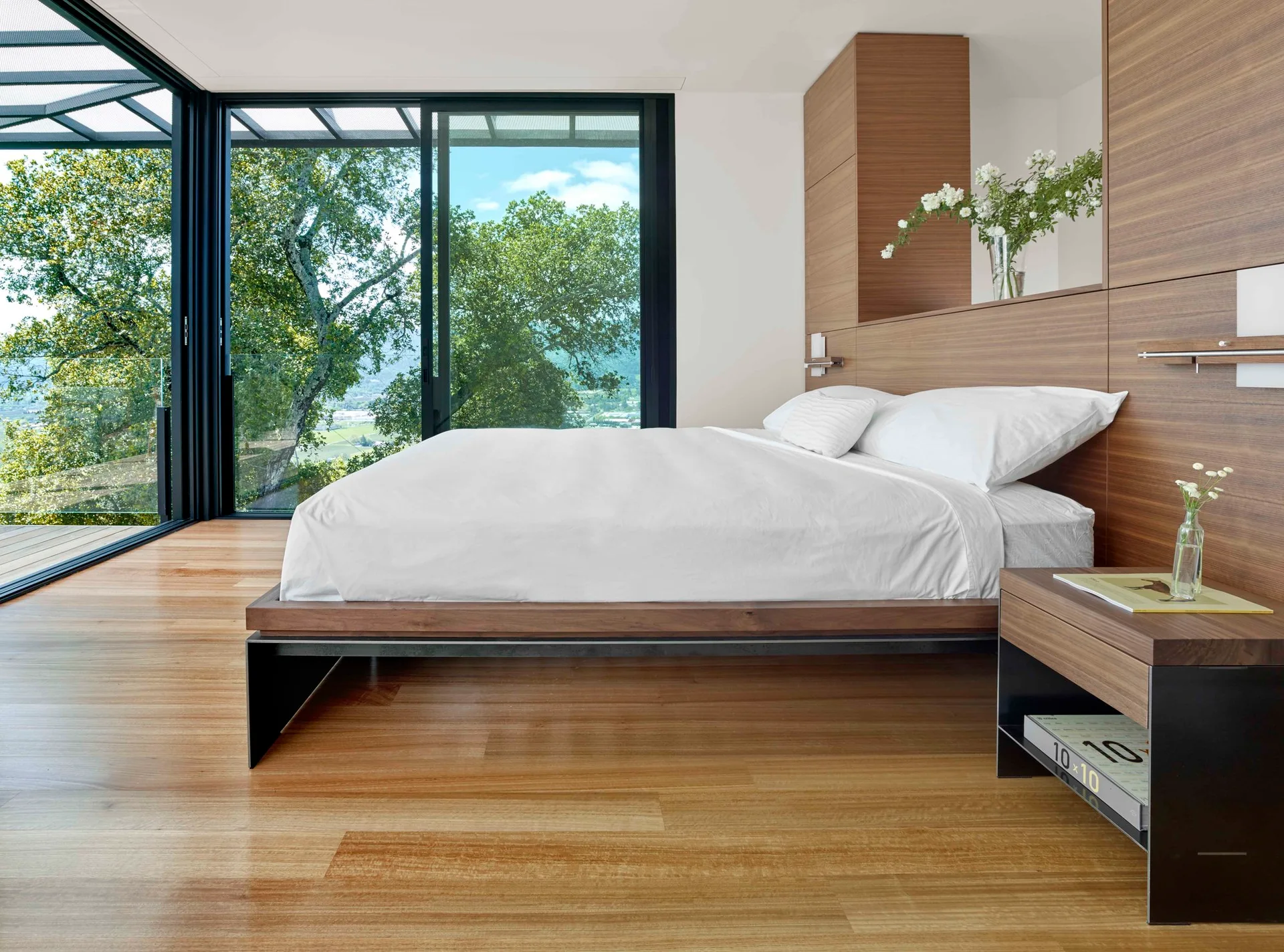 A modern minimalist bedroom featuring a low wood platform bed, an integrated wood panel headboard wall, and floor-to-ceiling glass doors opening to trees.