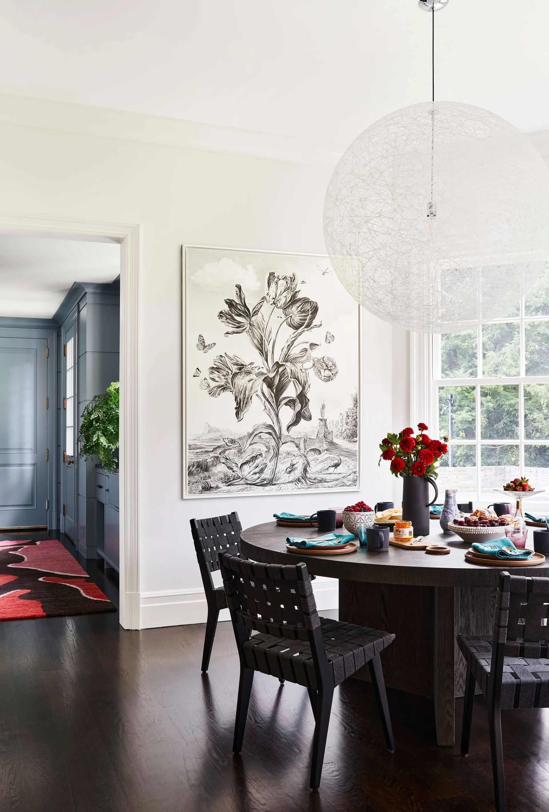 A round wooden dining table with woven strap chairs sits under a large sphere pendant light next to a framed black and white botanical print.