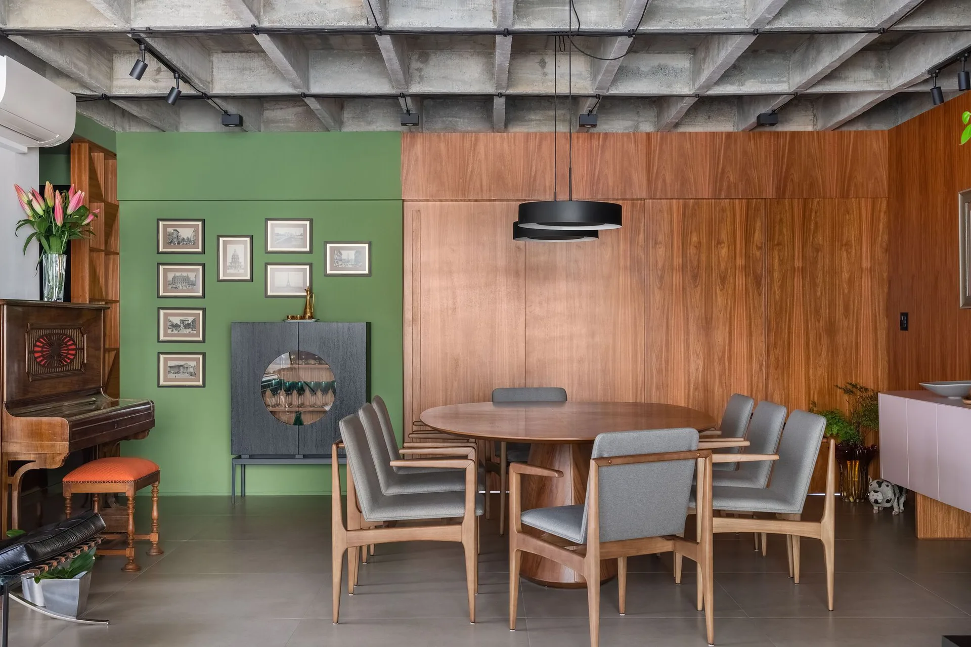 A modern dining room features a round wood table, gray chairs, and a split green and wood paneled wall below an exposed concrete ceiling.