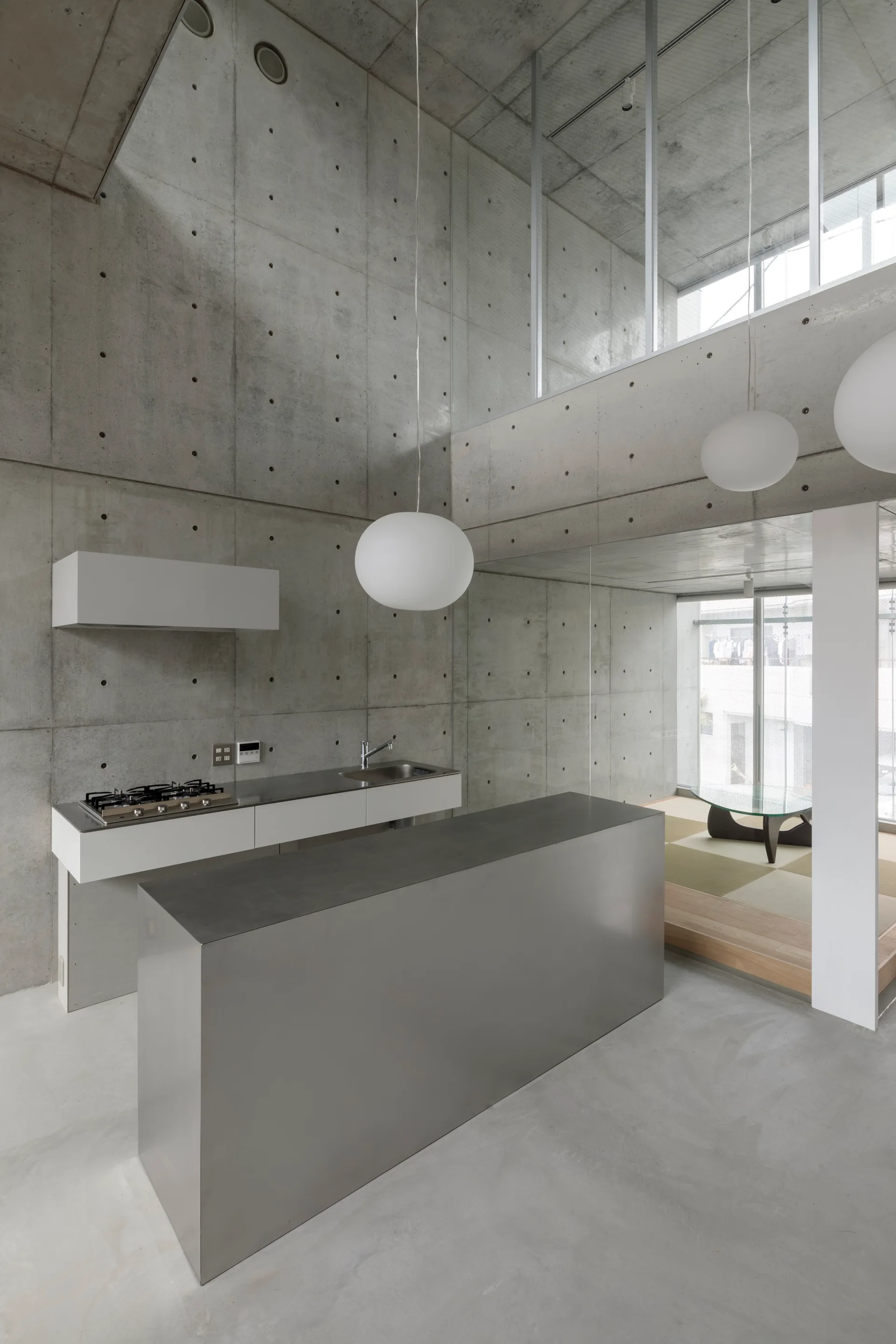 A minimalist kitchen features a metal island, a white counter, and globe pendant lights surrounded by double-height exposed concrete walls.