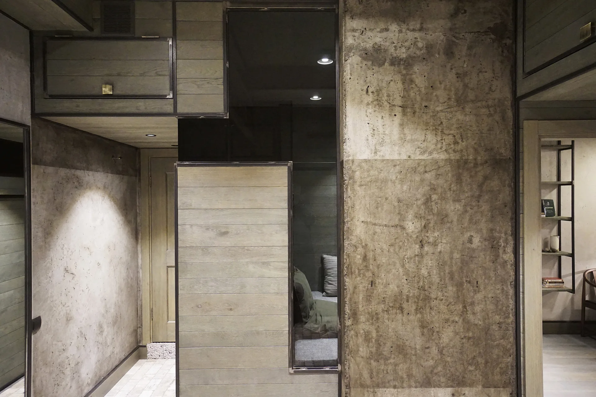 An interior hallway wall displays a mixture of rough exposed concrete and light wood paneling framed in black metal trim under recessed lights.