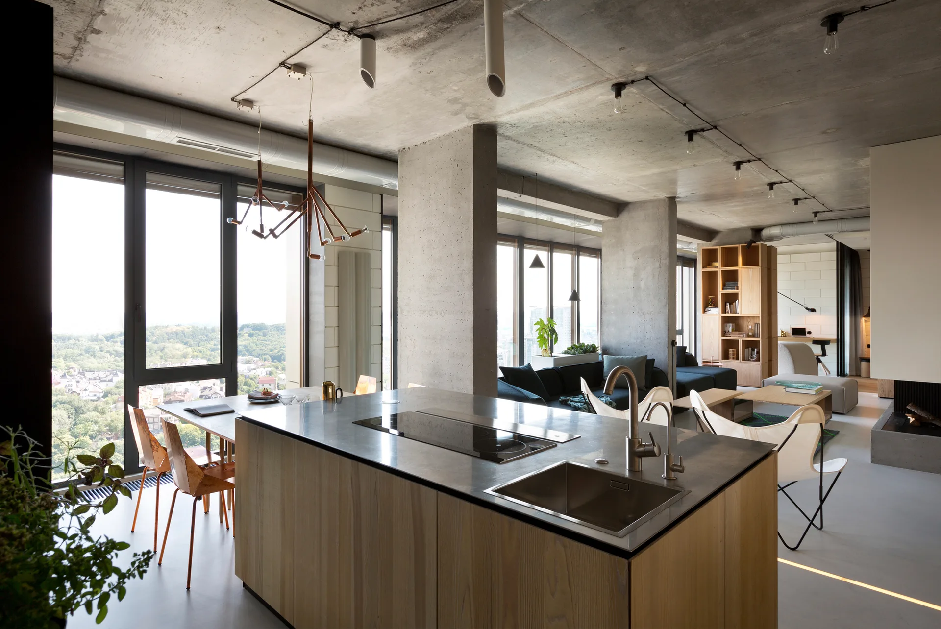 An open loft interior displays a wooden kitchen island under an exposed concrete ceiling, with concrete pillars separating the kitchen and living areas.
