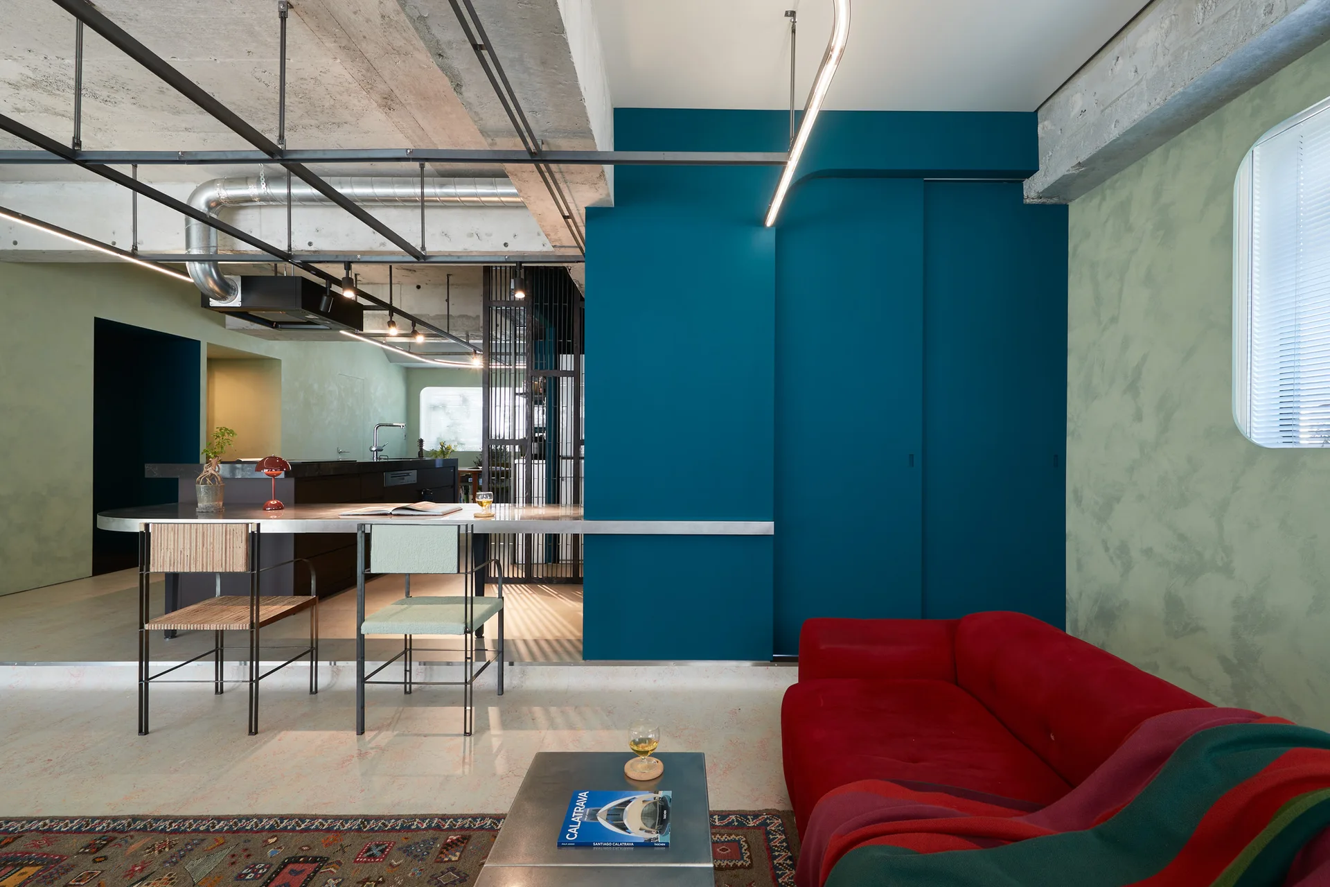 An industrial living room featuring a bright red sofa, a bold teal sliding partition wall, and an exposed raw concrete ceiling.