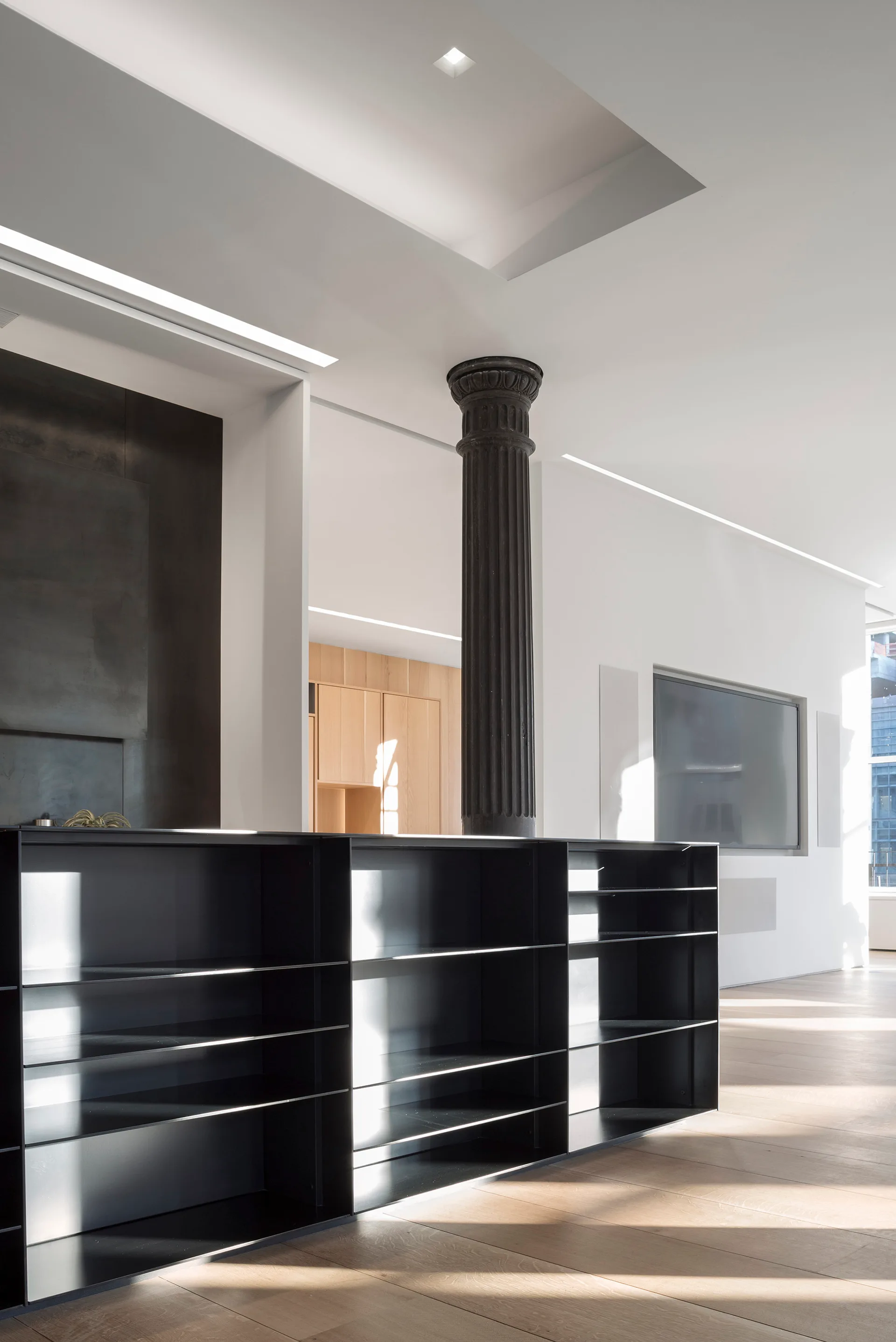 A minimalist white room features a dark fluted column, a low black metal shelving unit, and light wood floors.