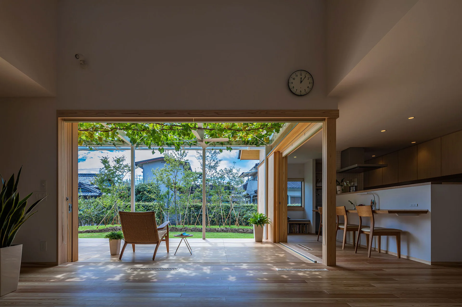A minimalist wood-paneled room opens to an outdoor deck with a wicker chair, small side table, and a green vine-covered trellis overhead.