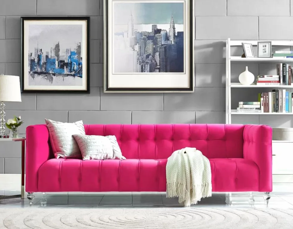 A modern living room features a bright pink tufted sofa with clear legs, set against a grey paneled wall decorated with framed art.